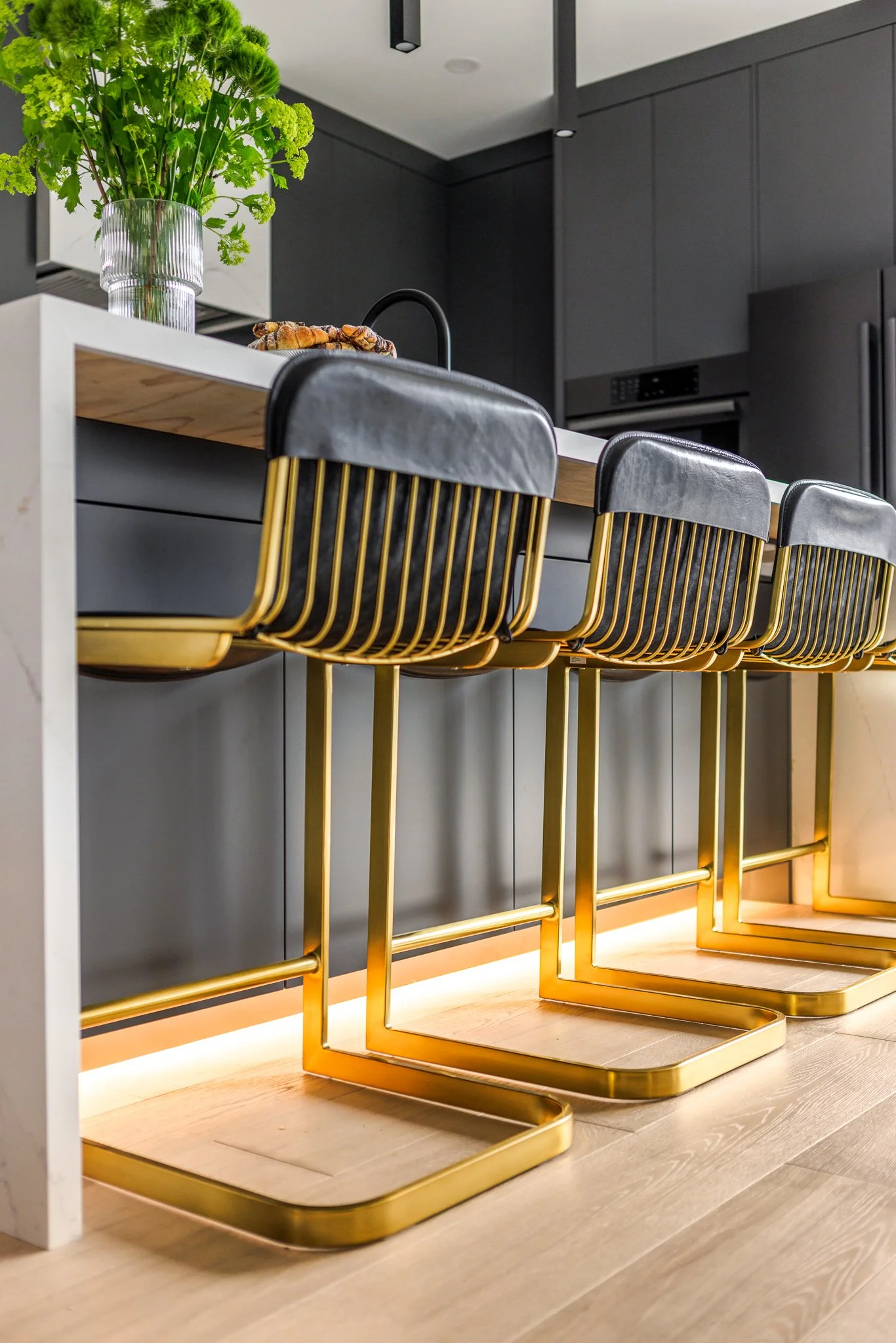 Modern kitchen with black cabinets, white countertop, green plant in glass vase, and three black and gold bar stools with wooden feet.