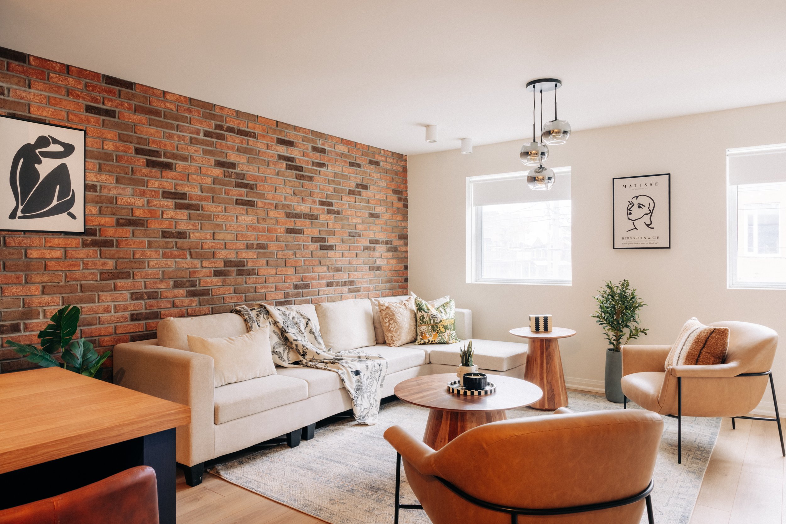 Cozy living room with a white sofa, beige armchairs, wooden coffee tables, a red brick accent wall, framed artwork, and potted plants, illuminated by natural light from two windows.