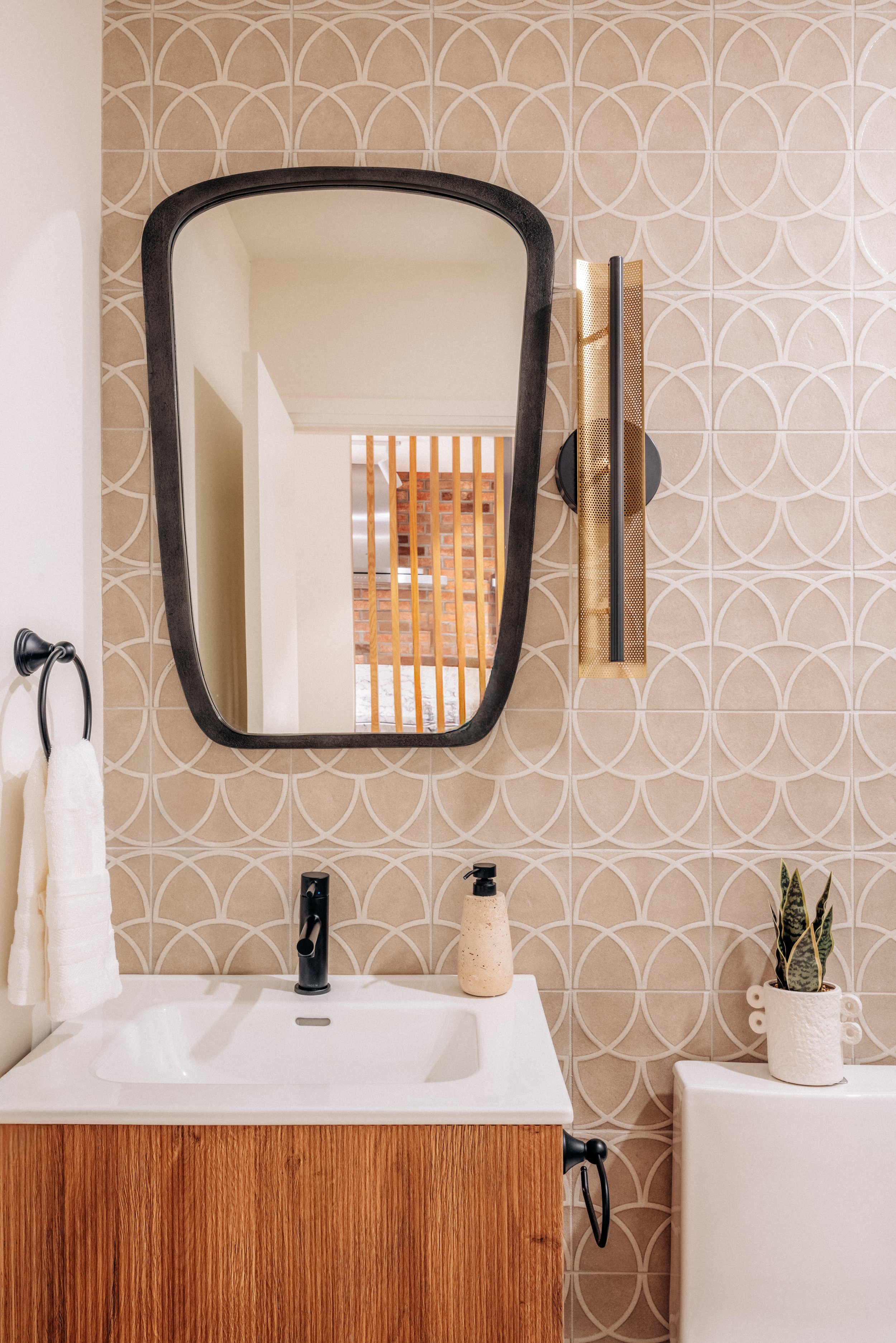 Modern bathroom with a frameless mirror, patterned beige tiles, a white sink with a wooden vanity, a black faucet, a soap pump, a potted snake plant, wall-mounted light fixture, and a toilet.