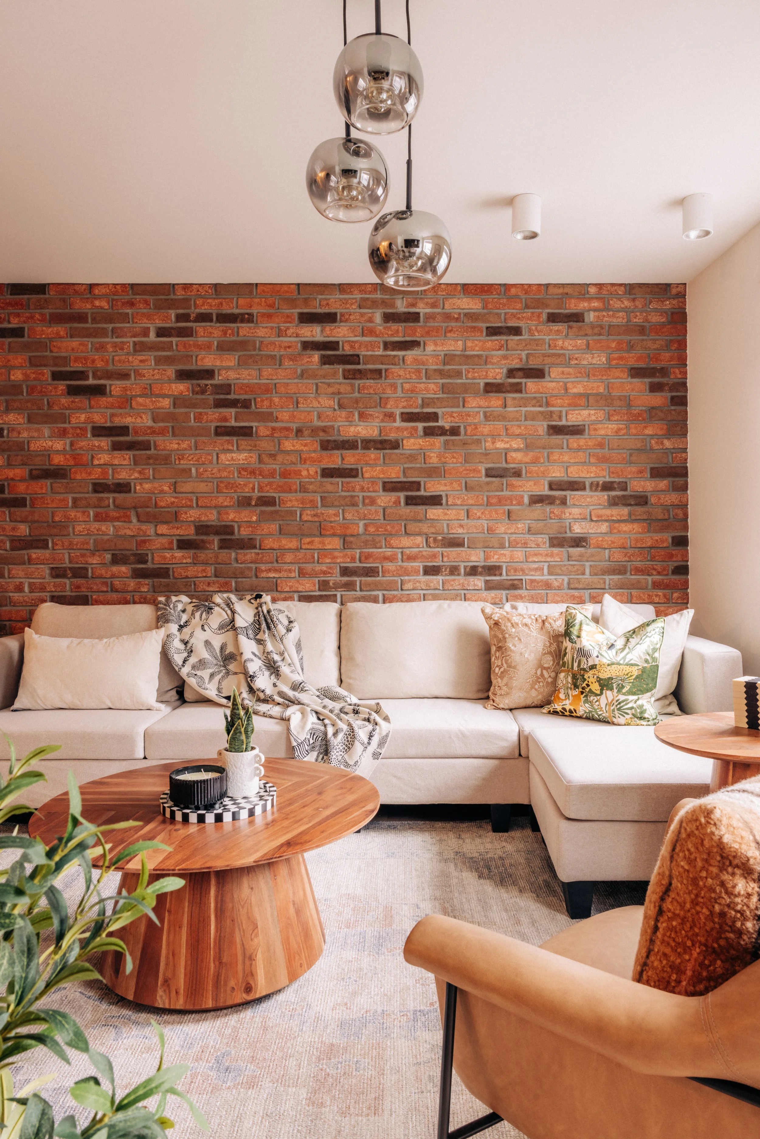Living room with a beige sectional couch, decorative pillows, a throw blanket, a round wooden coffee table with a candle and plant, a brick accent wall, and modern ceiling lighting fixtures.