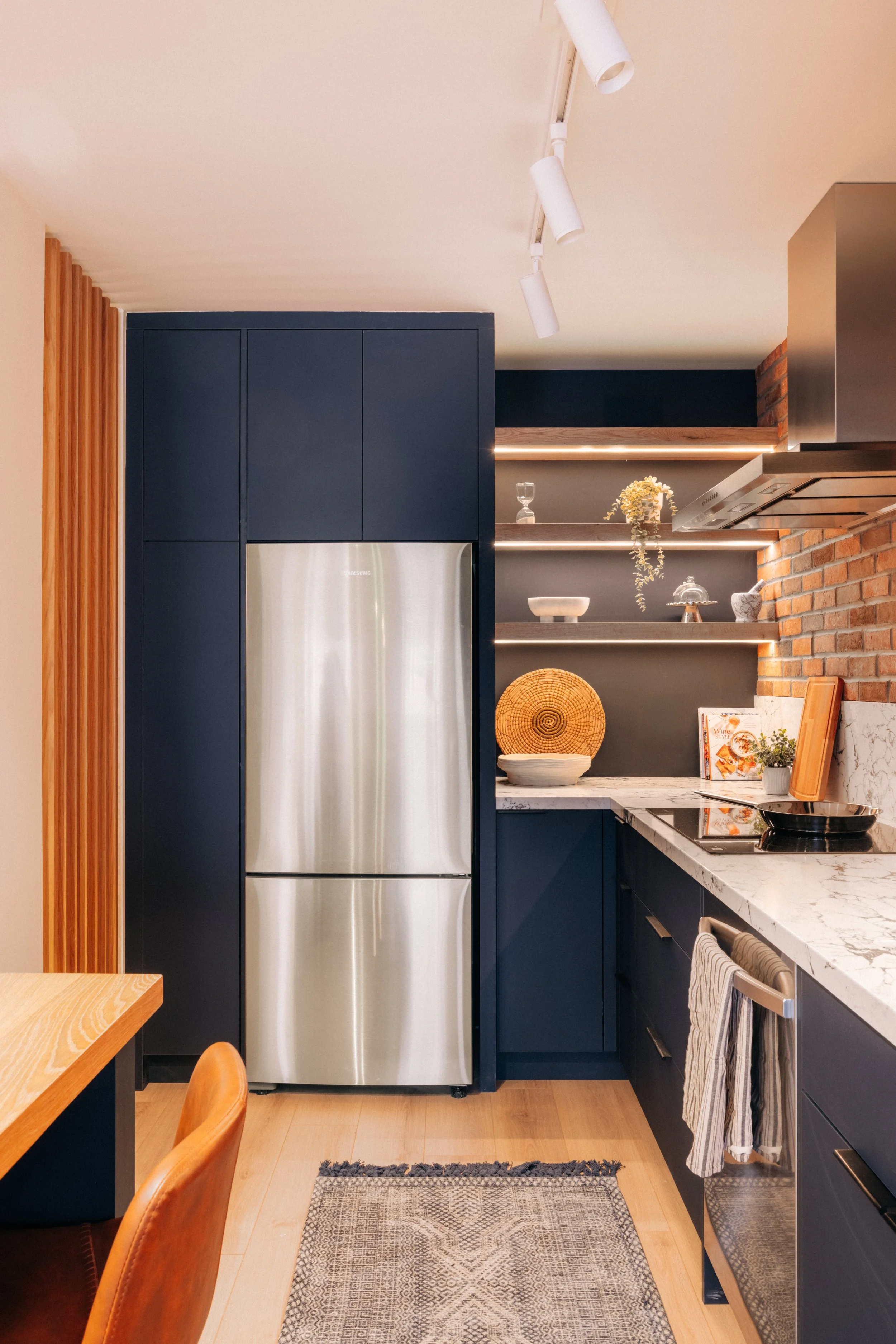 Modern kitchen with a stainless steel refrigerator, navy blue cabinets, open floating shelves with decor and cookbooks, marble countertop, and brick wall accents.