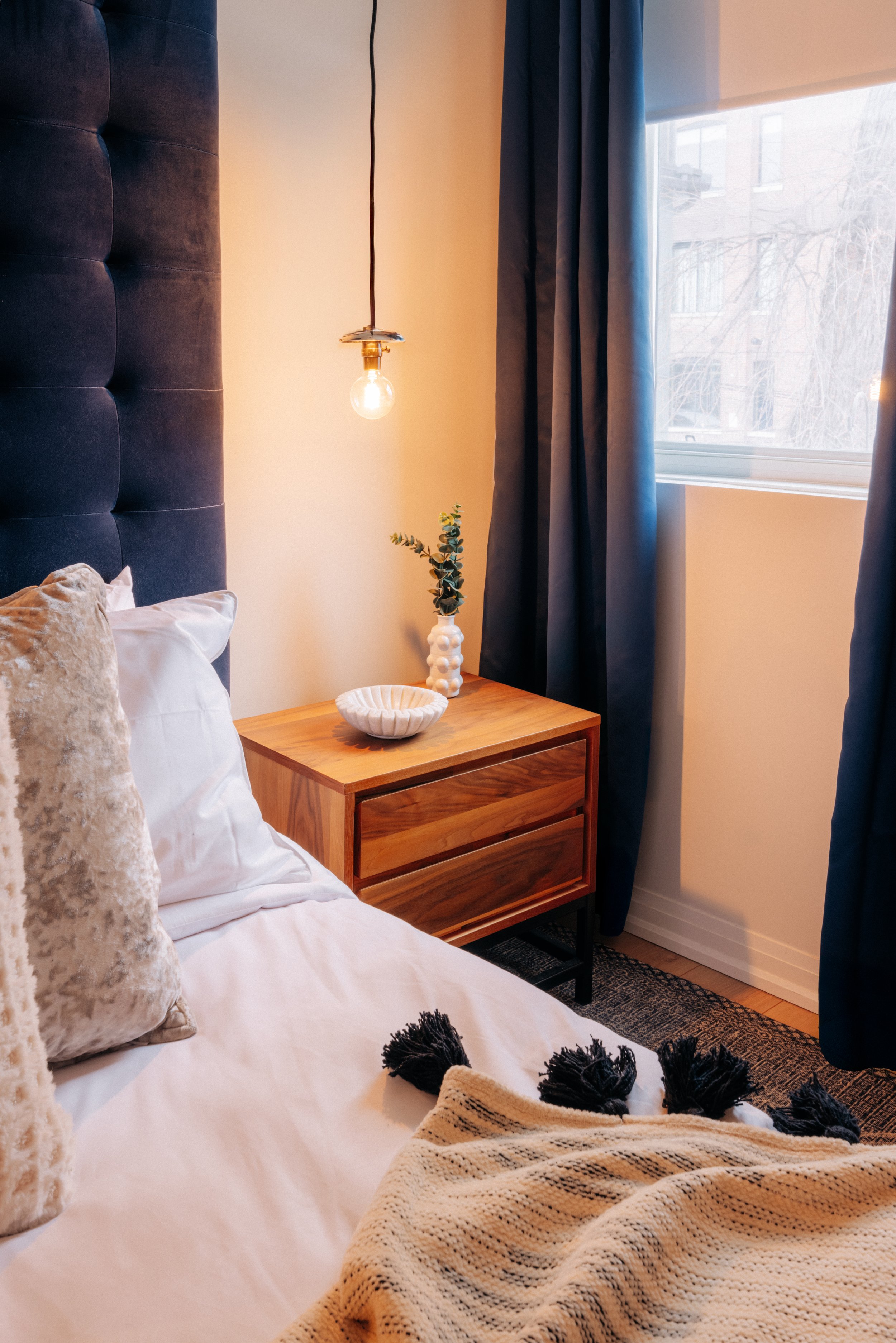 A cozy bedroom corner featuring a bed with cream and beige pillows, a beige throw blanket, a wooden nightstand with a white bowl and a small vase with green foliage, a hanging light bulb, dark blue curtains, and a window with a view of building and t