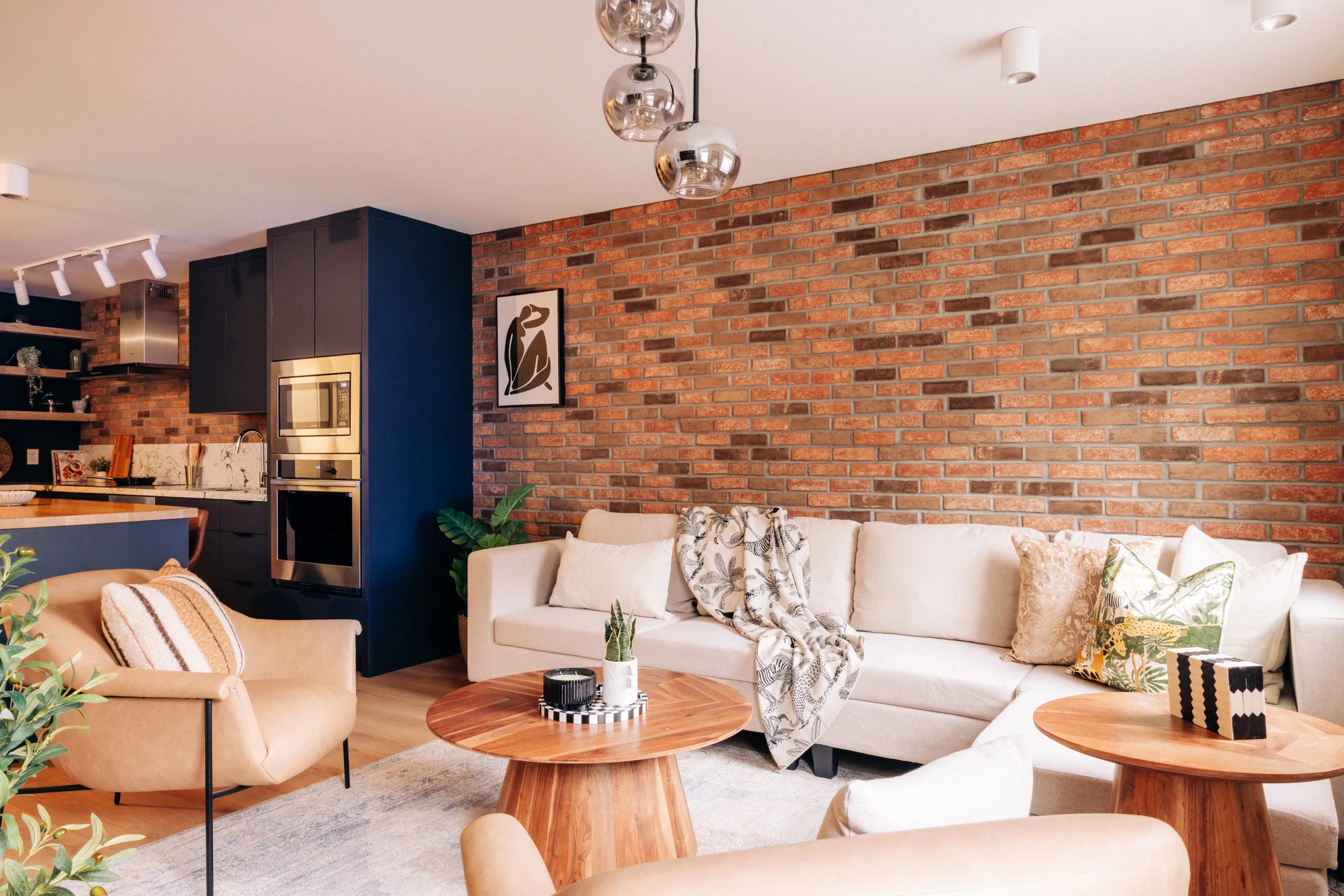 Living room with a beige sectional sofa, patterned pillows, and wooden coffee tables, accented by a brick wall, a beige armchair, and modern light fixtures.