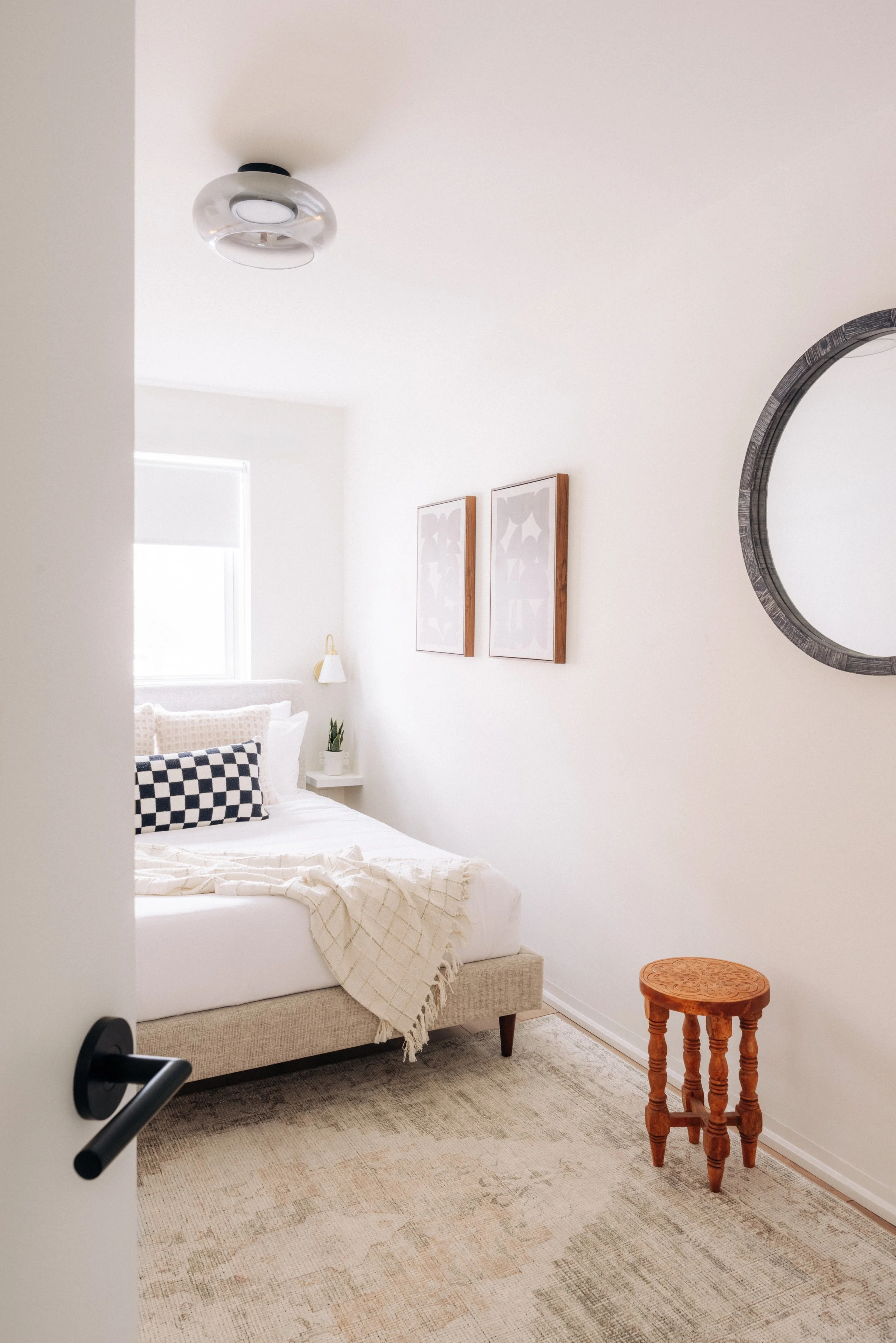 A bedroom with a beige bed, white linens, a checkered pillow, and a blanket, next to a window with a white shade, a small white nightstand with a plant, and wall art. There is a round mirror on the wall and a wooden stool on a light-colored rug.