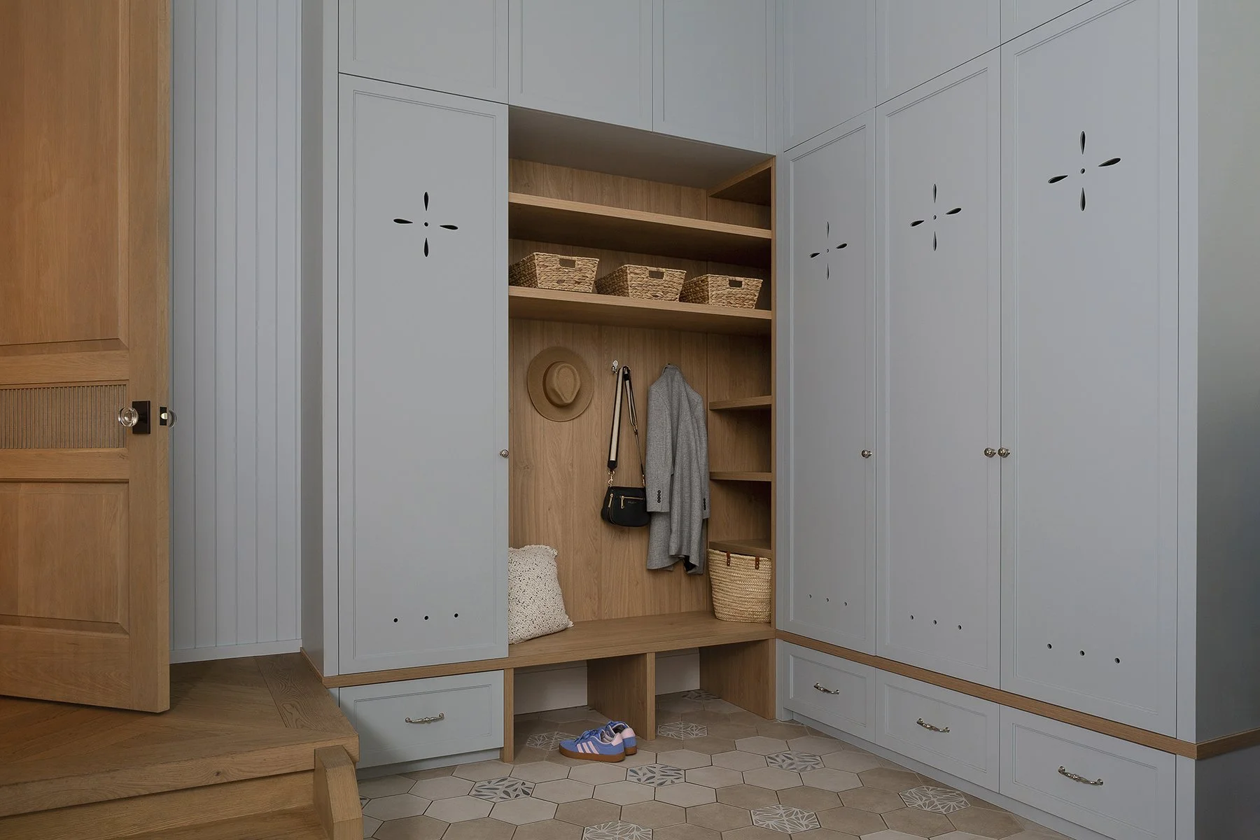 Stylish mudroom with light blue and natural wood cabinetry, open shelves with baskets, coat hooks with jacket, woven basket, colorful shoes, and patterned tile flooring.