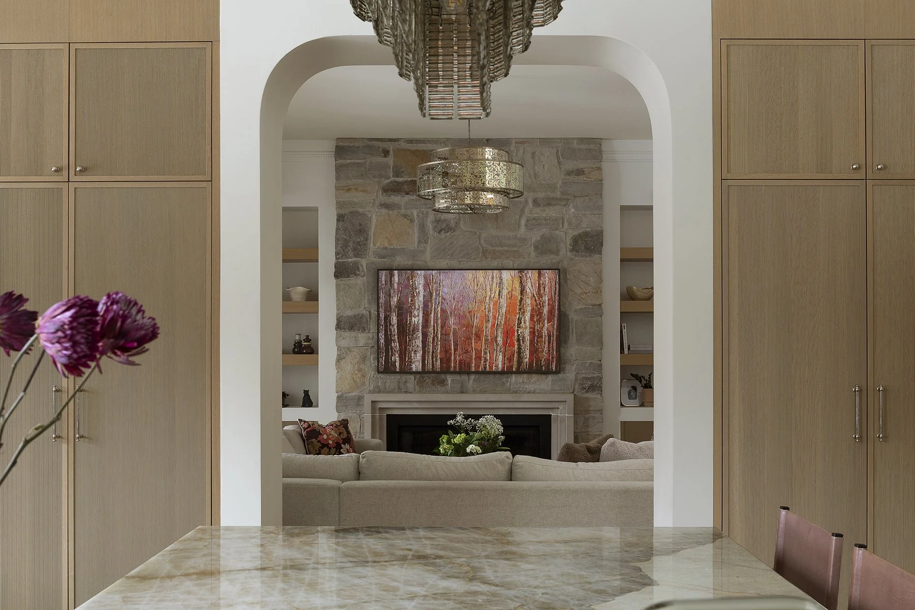 View through an archway into a living room with a stone fireplace, a wall-mounted television showing colorful autumn trees, a beige sectional sofa with decorative pillows, and built-in shelves with decor items. Modern chandelier hangs from the ceilin