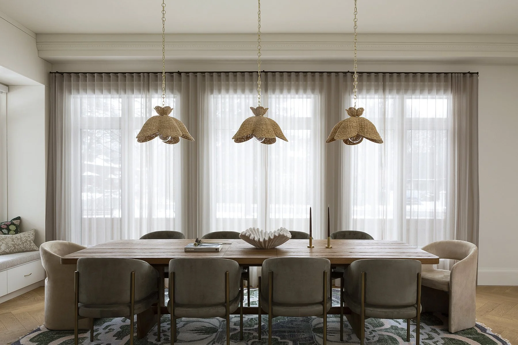 Dining room with a wooden table, eight chairs, three hanging wicker lamps, large windows with sheer curtains, and decorative items on the table.