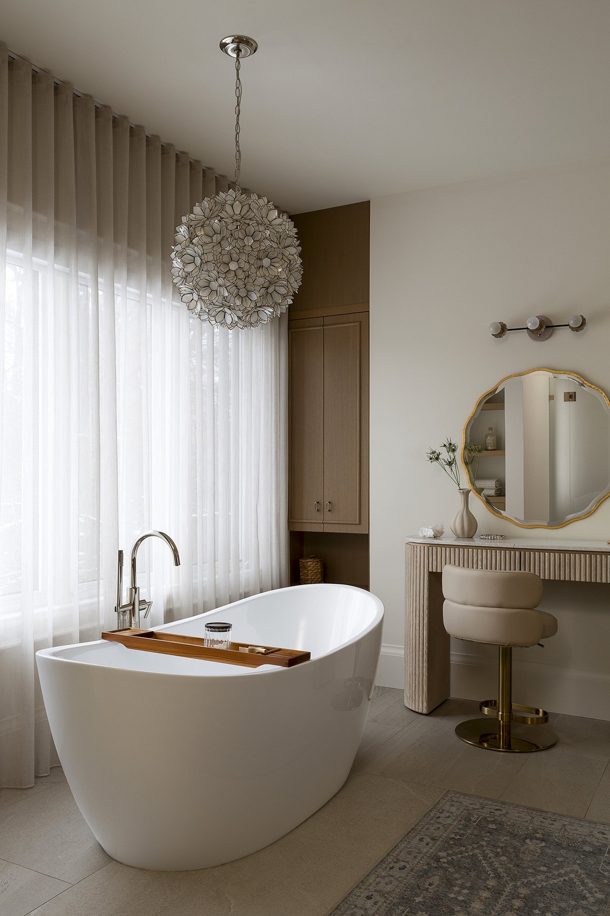 Modern bathroom with a freestanding white bathtub, a wooden tray, sheer curtains, and a vanity with a mirror and chair.