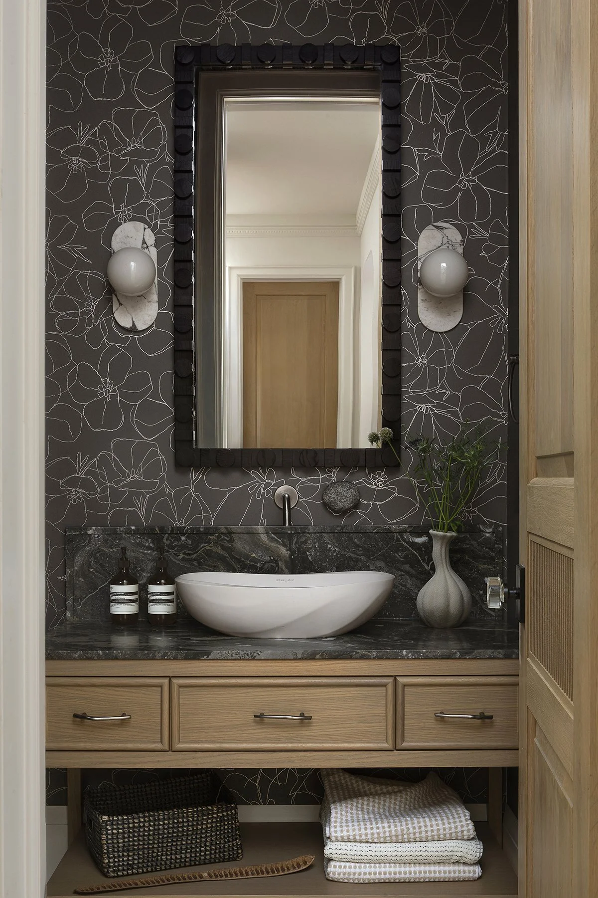 A modern bathroom vanity with a dark marble countertop, white vessel sink, large mirror with black framed border, two wall-mounted light fixtures, a beige wooden cabinet, and decorative items including two brown bottles, a gray vase with green plants