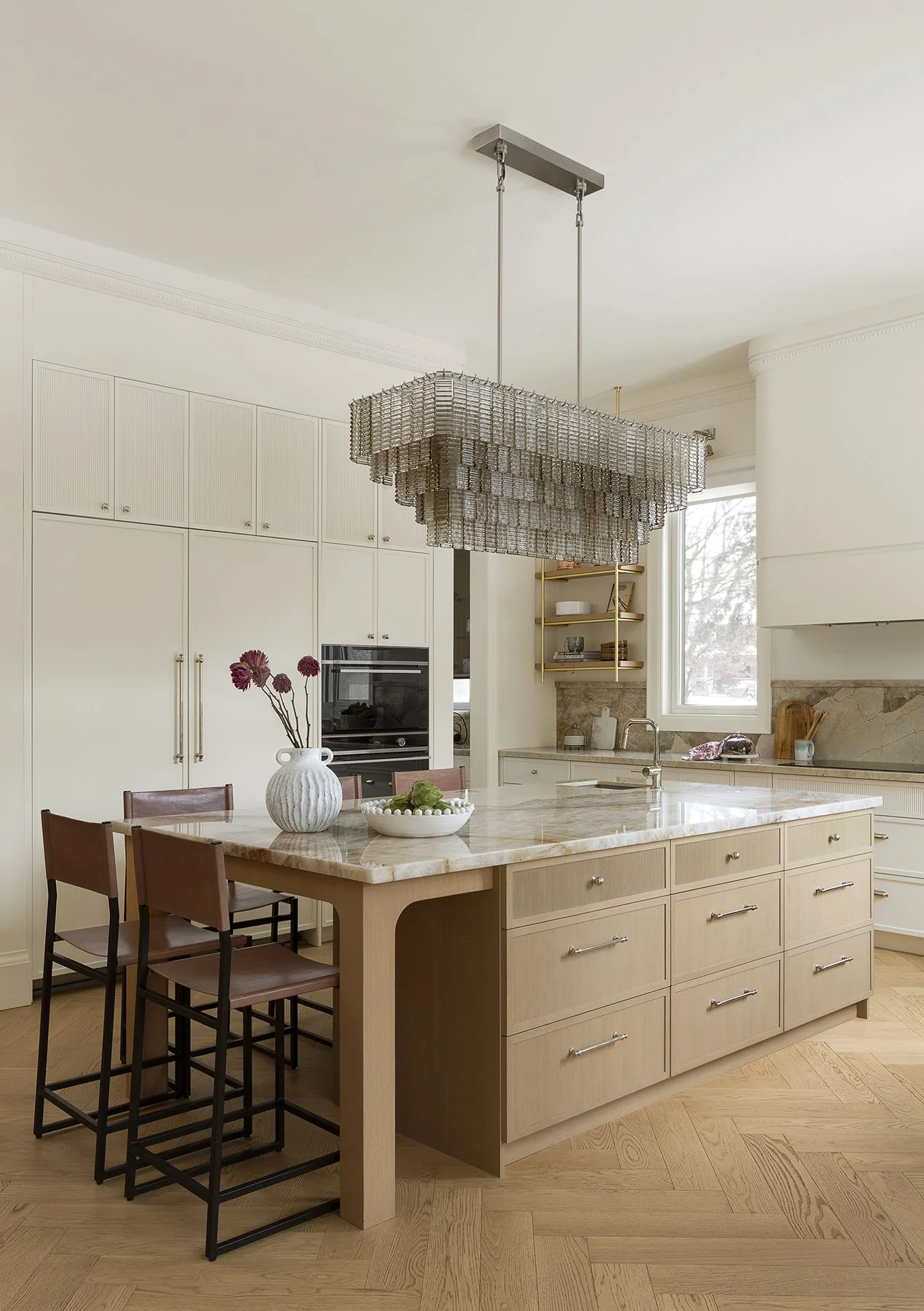 Modern kitchen with beige and white cabinetry, a large marble island with five chairs, a decorative chandelier overhead, and open shelves with dishes near a window.