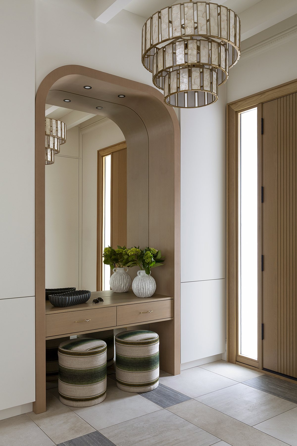 Entryway with a wooden and white cabinet, two striped round ottomans, a large mirror, potted plants, and modern chandeliers.