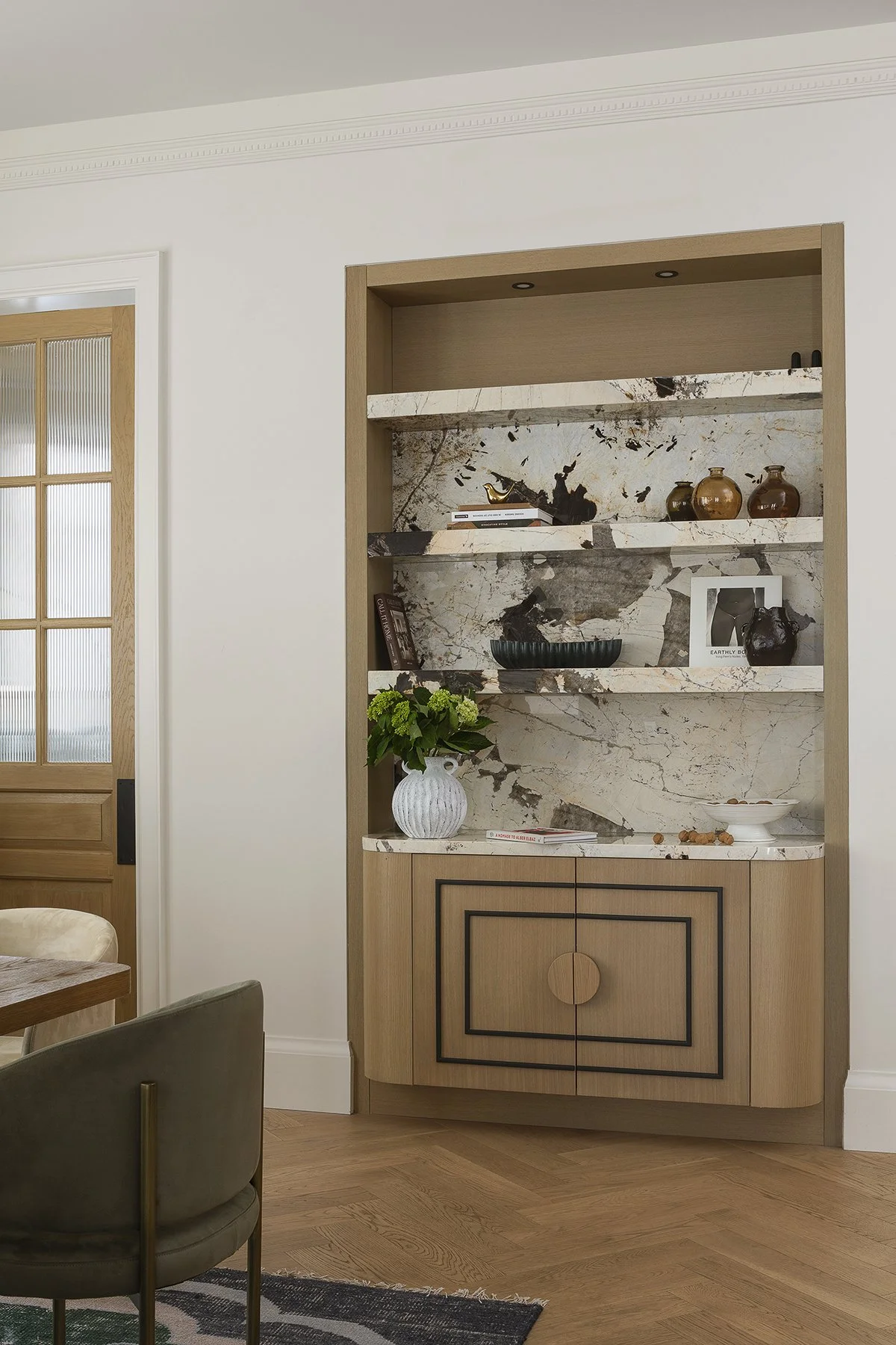 A wooden and marble open shelf unit in a room with a wooden floor and white walls, decorated with vases, books, framed pictures, a bowl, and a vase with green flowers.