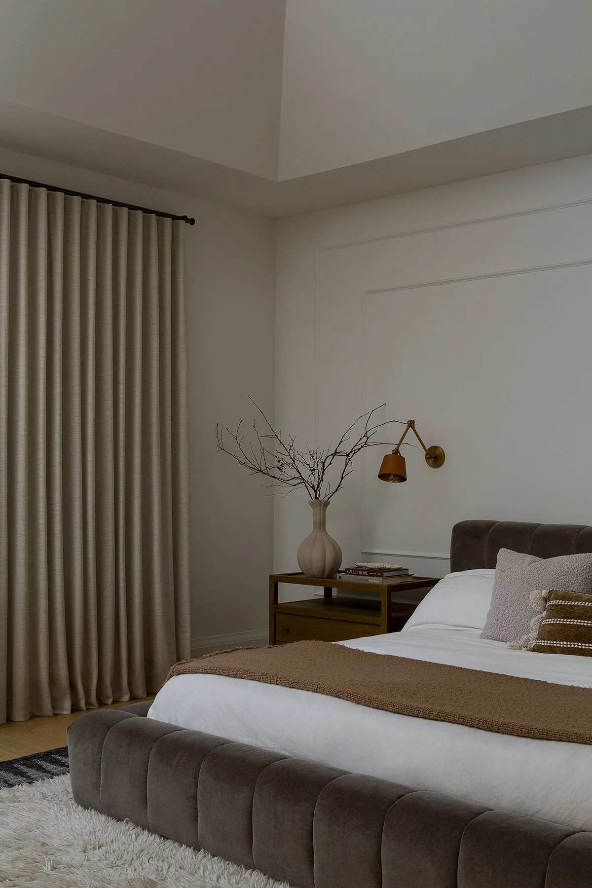 A bedroom with a bed, side table, and beige curtains, decorated with a vase with branches and a wall-mounted reading lamp.