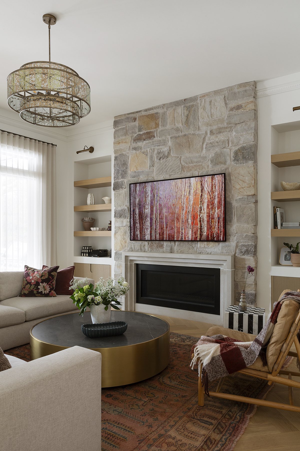 Living room with a stone fireplace, wall-mounted TV, beige sofa with floral pillows, a round coffee table with a black and gold finish, a flower vase, various shelves with decor, and a rocking chair with a throw blanket.