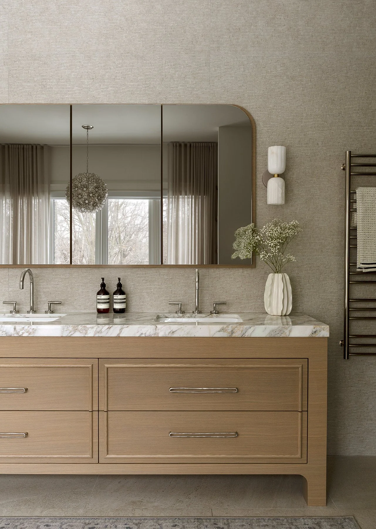 Bathroom vanity with a large mirror, two sinks, a marble countertop, two black bottles, a vase with flowers, and a wall-mounted towel rack.