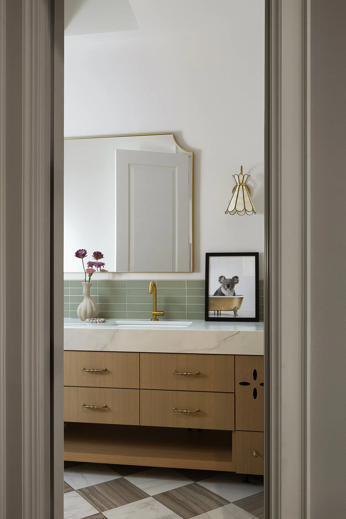A bathroom vanity with a marble countertop, green tile backsplash, and a large mirror. Decor includes a vase with purple flowers, a framed picture of a koala in a bathtub, and a wall sconce with a scalloped shade. The vanity has wood drawers with bra