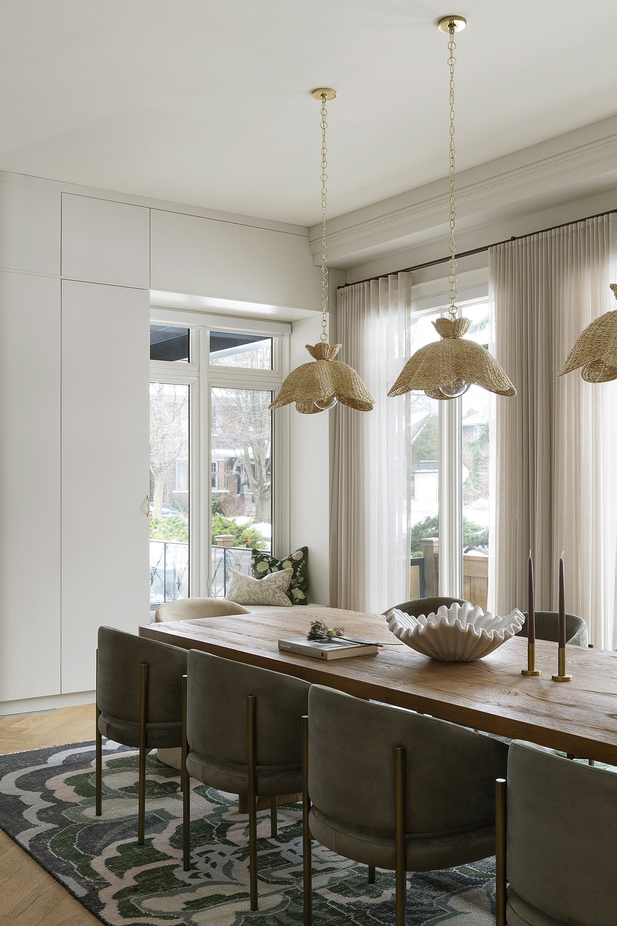 Dining room with a wooden table, six gray upholstered chairs, a white bowl centerpiece, and two tall candlesticks. Pendant lights with woven shades hang above. Large windows with sheer curtains let in natural light.