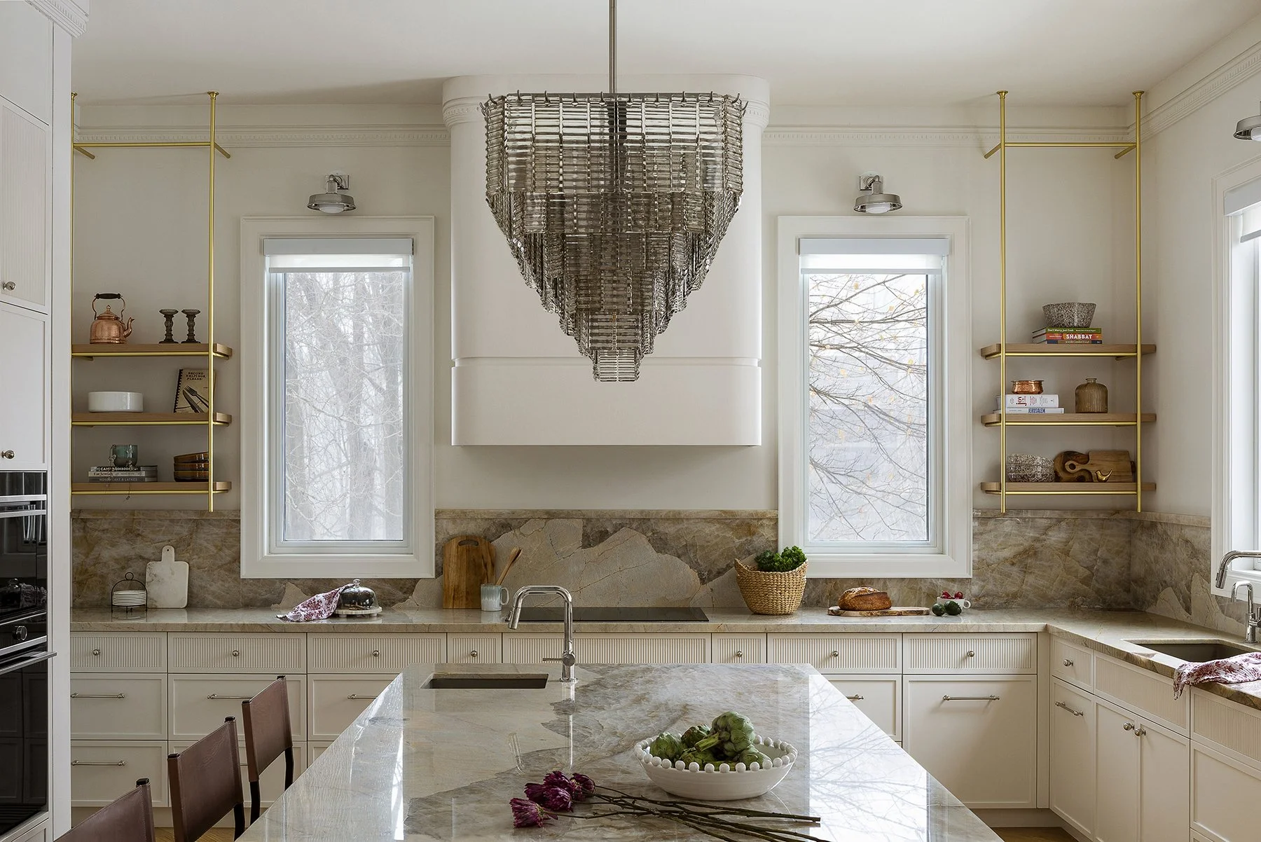 Modern kitchen with marble countertops, open shelving, and a large decorative chandelier hanging from the ceiling. Two large window provide natural light, and various decorative and kitchen items are placed throughout.