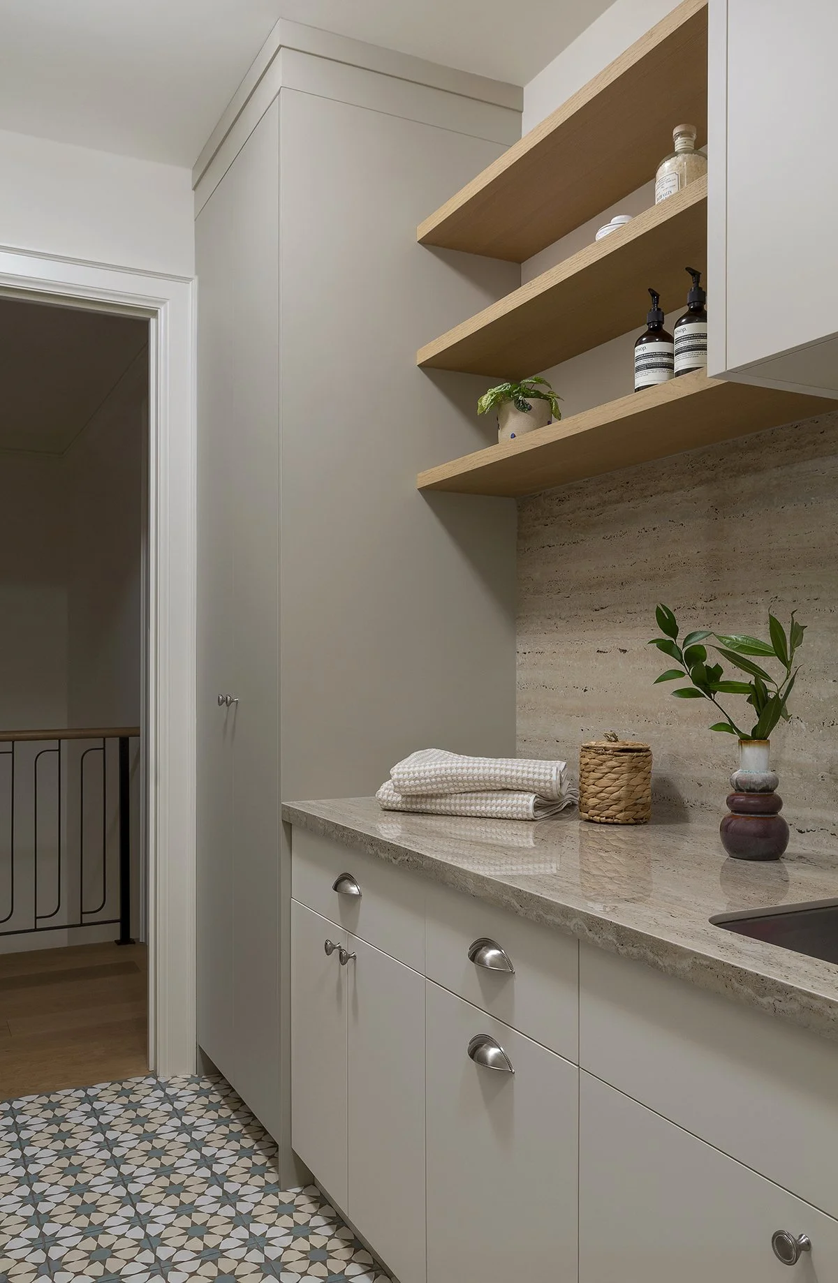 Modern kitchen with beige marble countertop, white cabinetry with metallic handles, open wooden shelves with bottles and jars, decorative vases with plants, patterned tiled floor, and a doorway leading to a wooden-floored area.