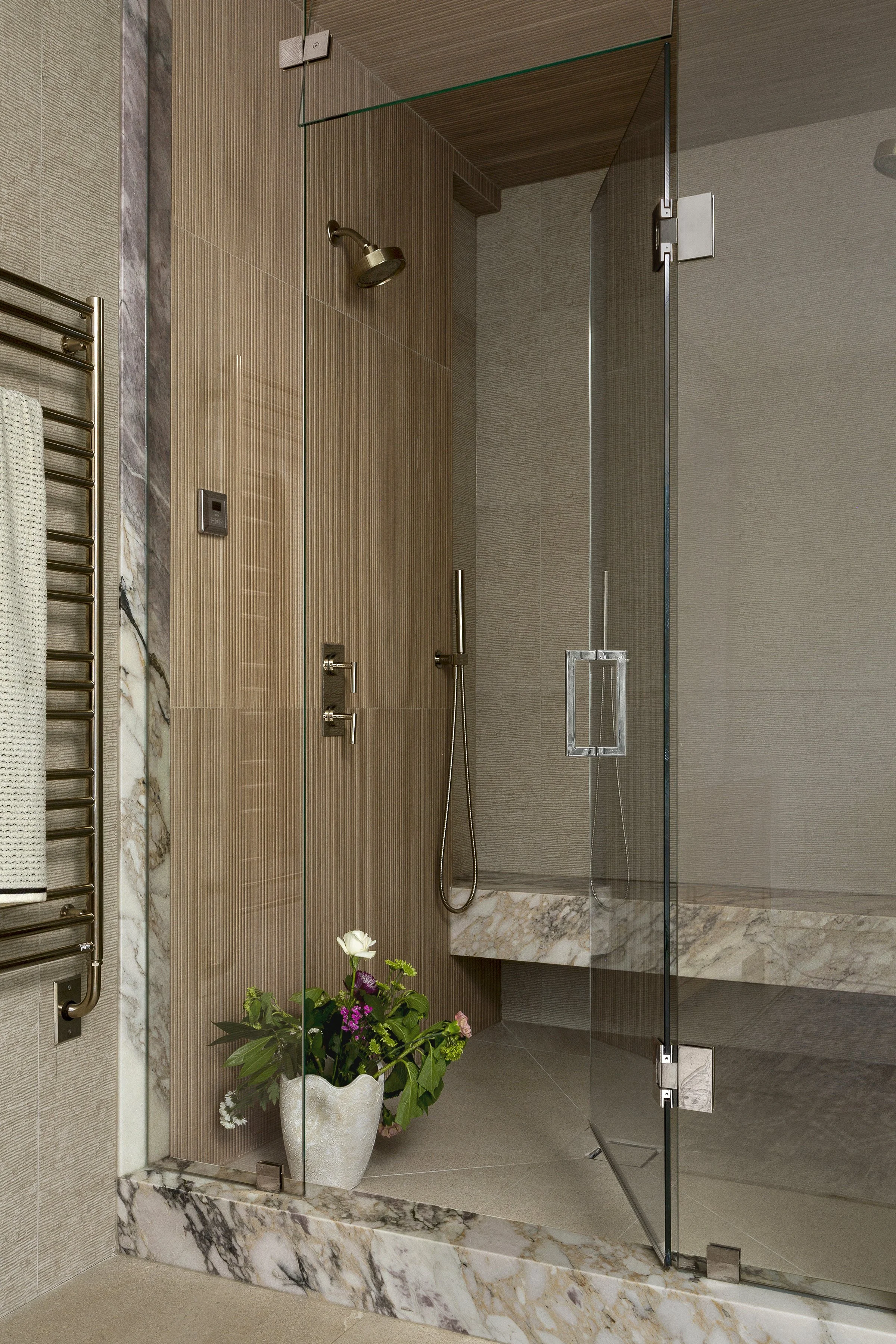 Modern walk-in shower enclosed with clear glass, featuring a built-in marble bench, a rainfall showerhead, a handheld shower, and a potted plant with flowers at the shower entrance.