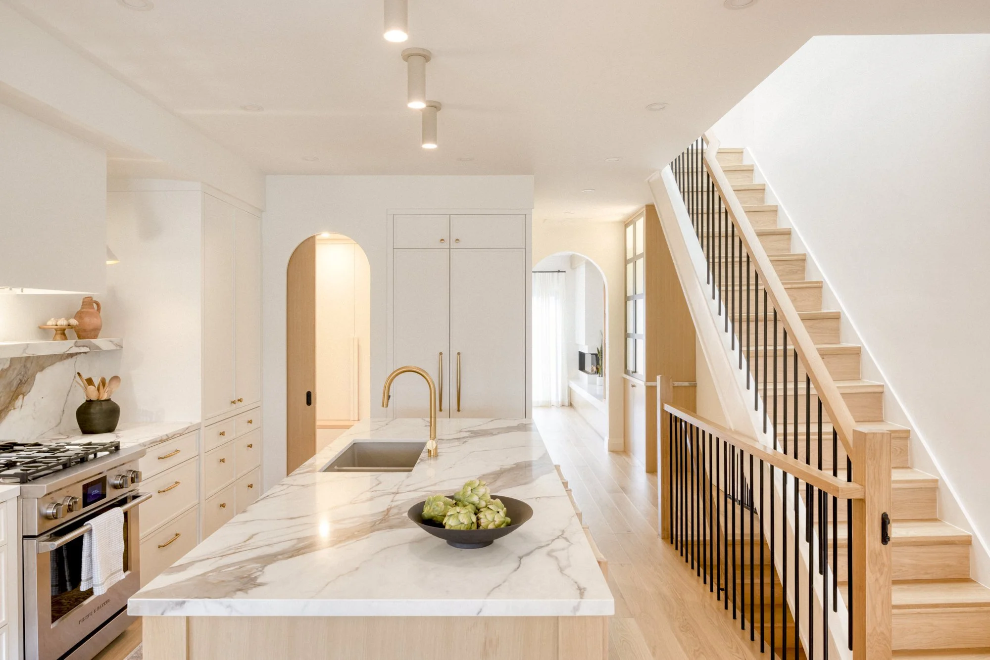 Modern kitchen with marble countertop island, white cabinets, stainless steel oven, wooden staircase with black railing, and natural light through large window.