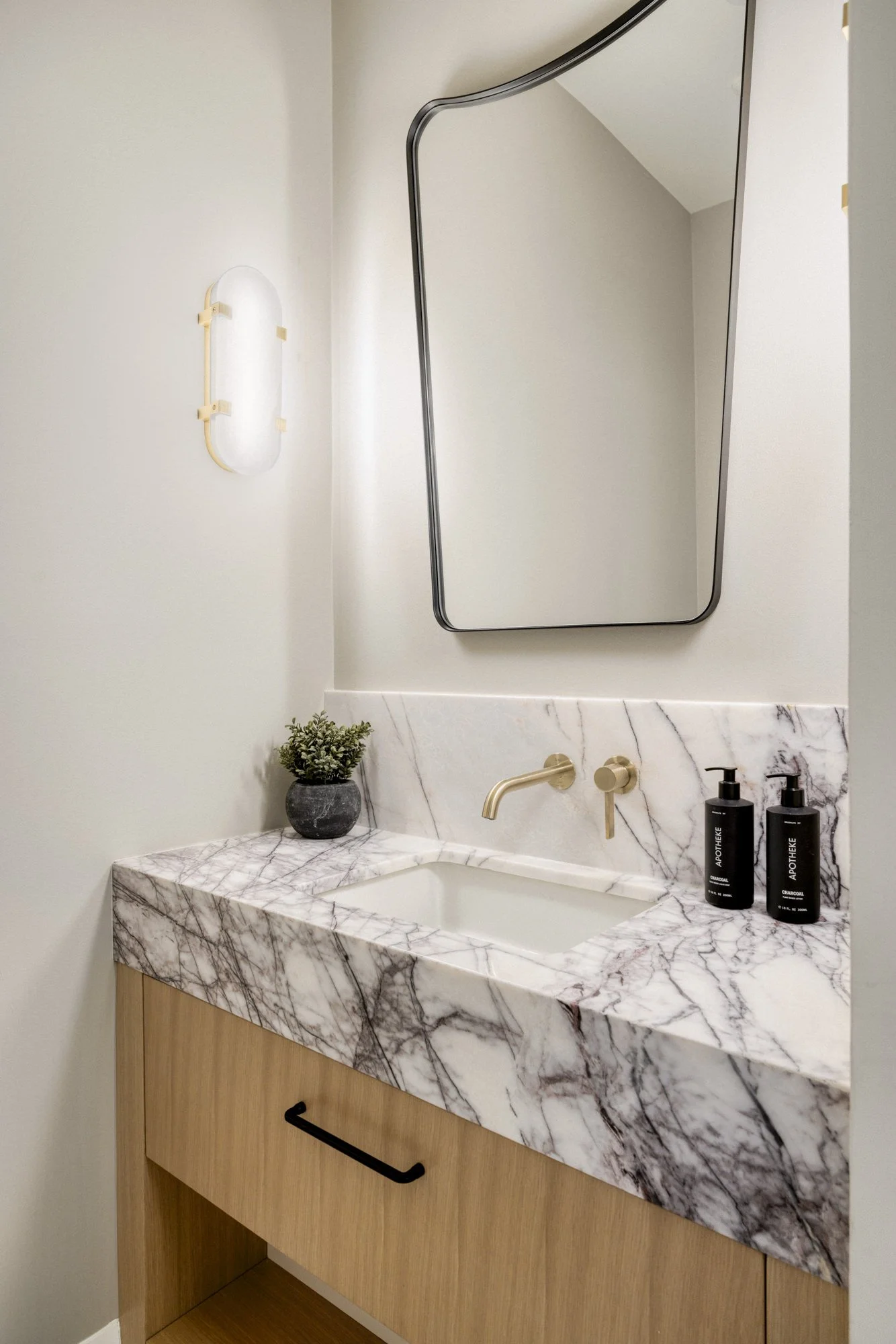Modern bathroom vanity with white and gray marble countertop, wooden cabinet, black handle, black soap dispenser, small potted plant, oval mirror, and wall-mounted faucet, with a wall light fixture.