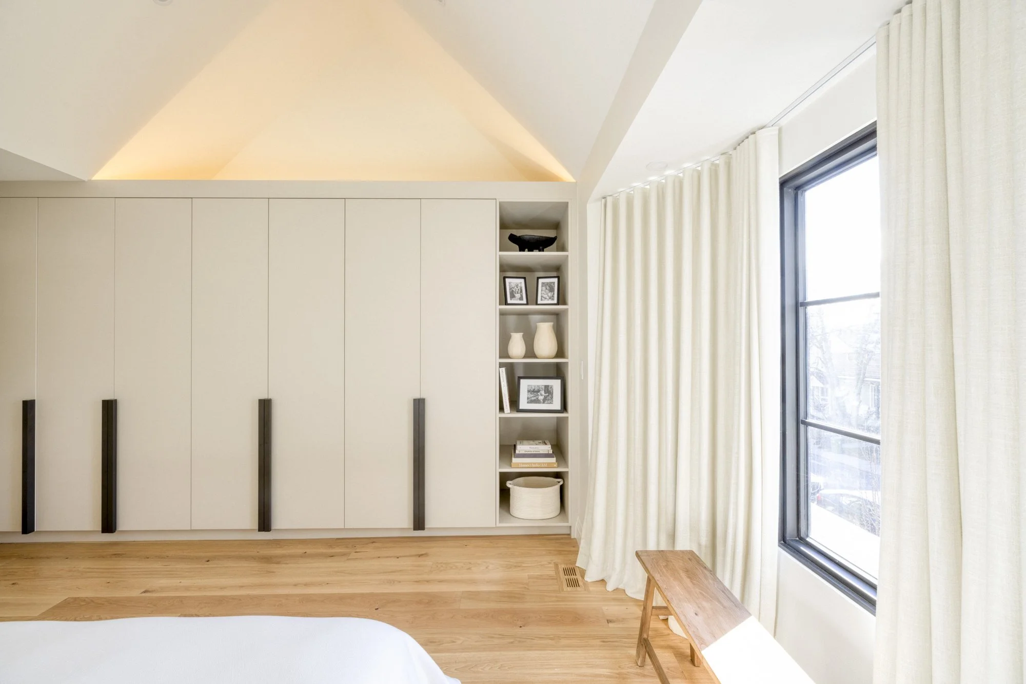 A mostly white bedroom with a large tall wardrobe, built-in open shelves with decor, a window with white curtains, and a small wooden bench by the window. The room has light wood flooring and a vaulted ceiling.