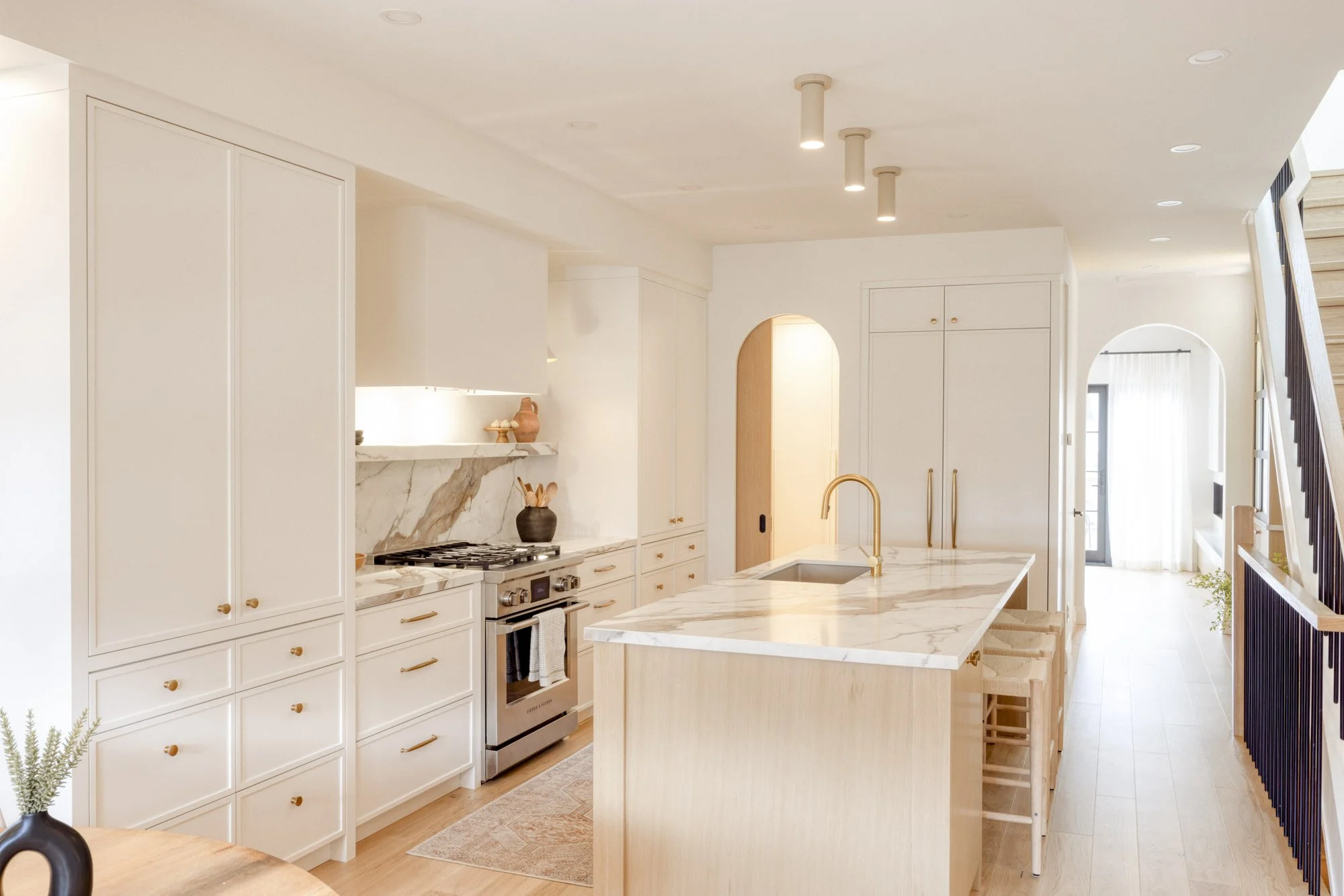 Modern white kitchen with marble countertops, built-in oven, and a large island with a gold faucet, light wood cabinets, and a staircase on the right let into a bright living space.