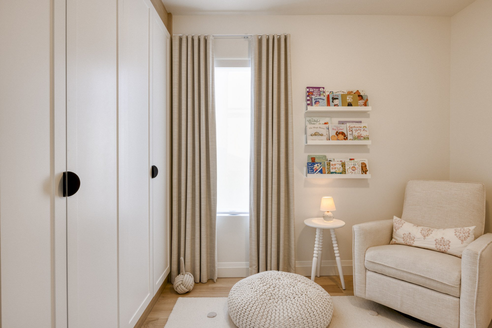 Bedroom with a white wardrobe, beige curtains, small white side table with a lamp, cream armchair with a floral pillow, knitted pouf, white shelves with children's books, beige walls, and hardwood floor.