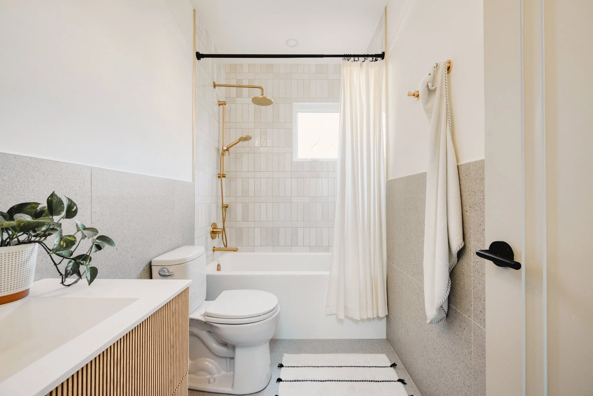 Modern bathroom with a bathtub and shower, gold fixtures, a window with a curtain, a white toilet, a small white and wood cabinet, a plant in a pot, and a striped rug.