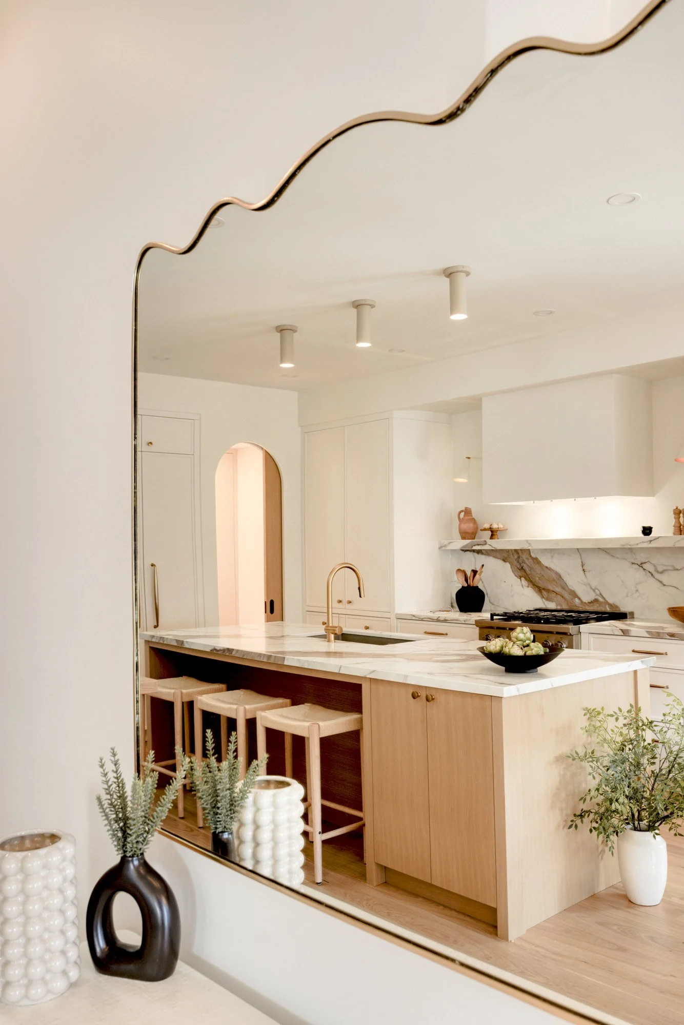 Modern kitchen with marble island, wooden cabinets, and gold faucet, seen through a large mirror with a gold wavy border. Decor includes vases with plants and pottery.