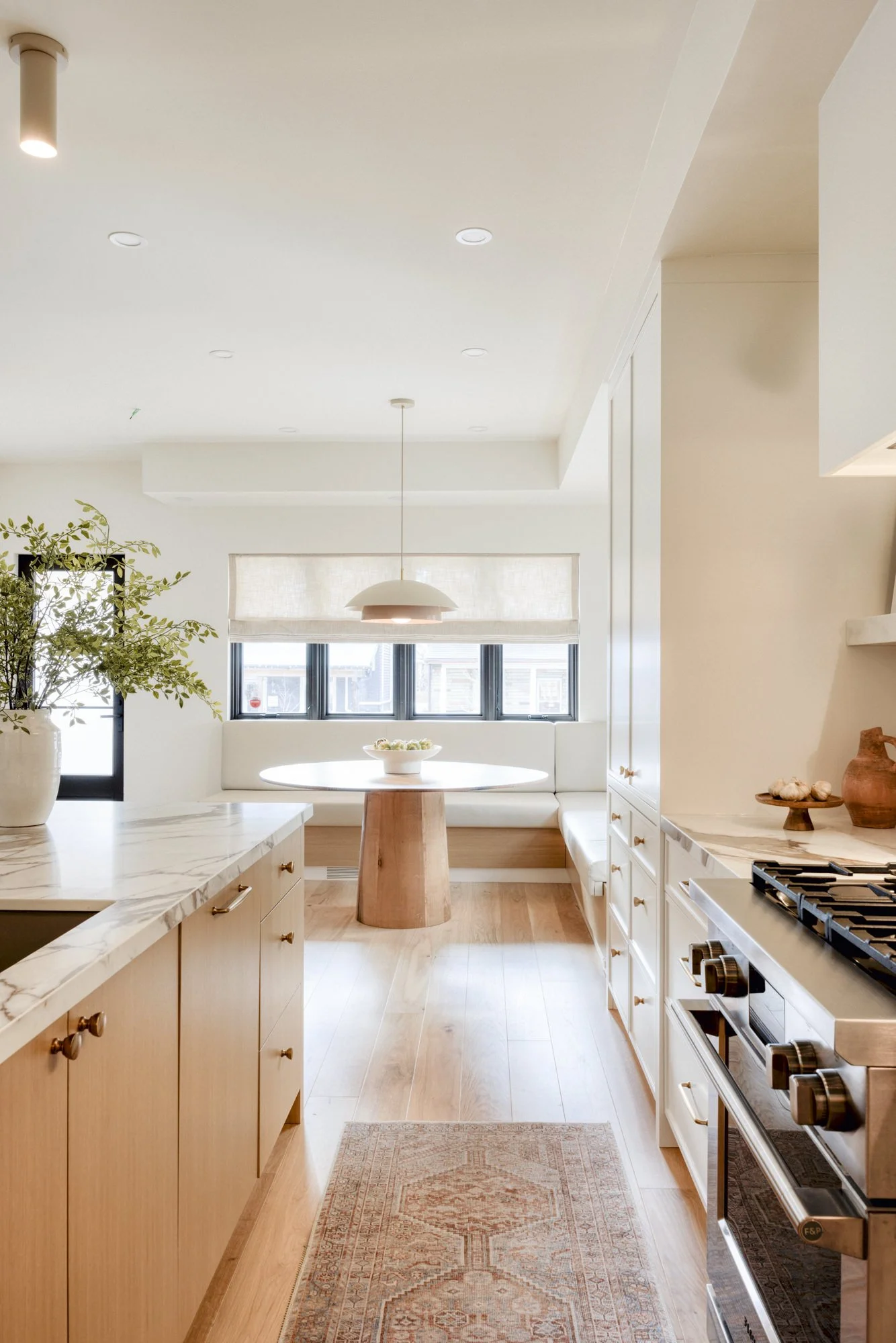 Bright kitchen with white cabinets, marble countertops, and stainless steel stove. A window seat with a round table and hanging lamp in the background. Green plant on the counter and decorative pottery on the side.