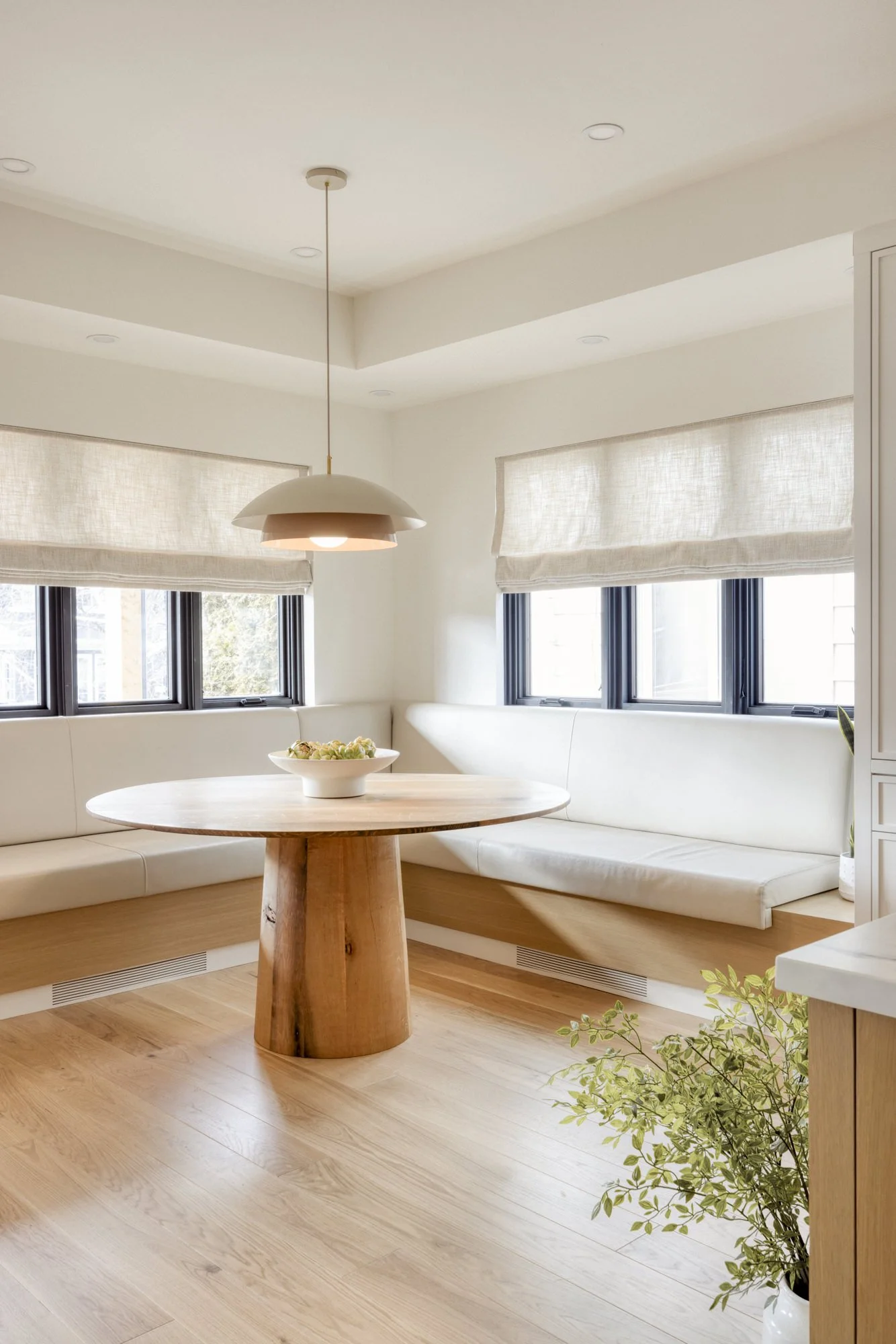 A cozy dining nook with a round wooden table and white cushioned bench seating, illuminated by a hanging pendant light. The space features light wood flooring, large windows with beige shades, and a small potted plant nearby.