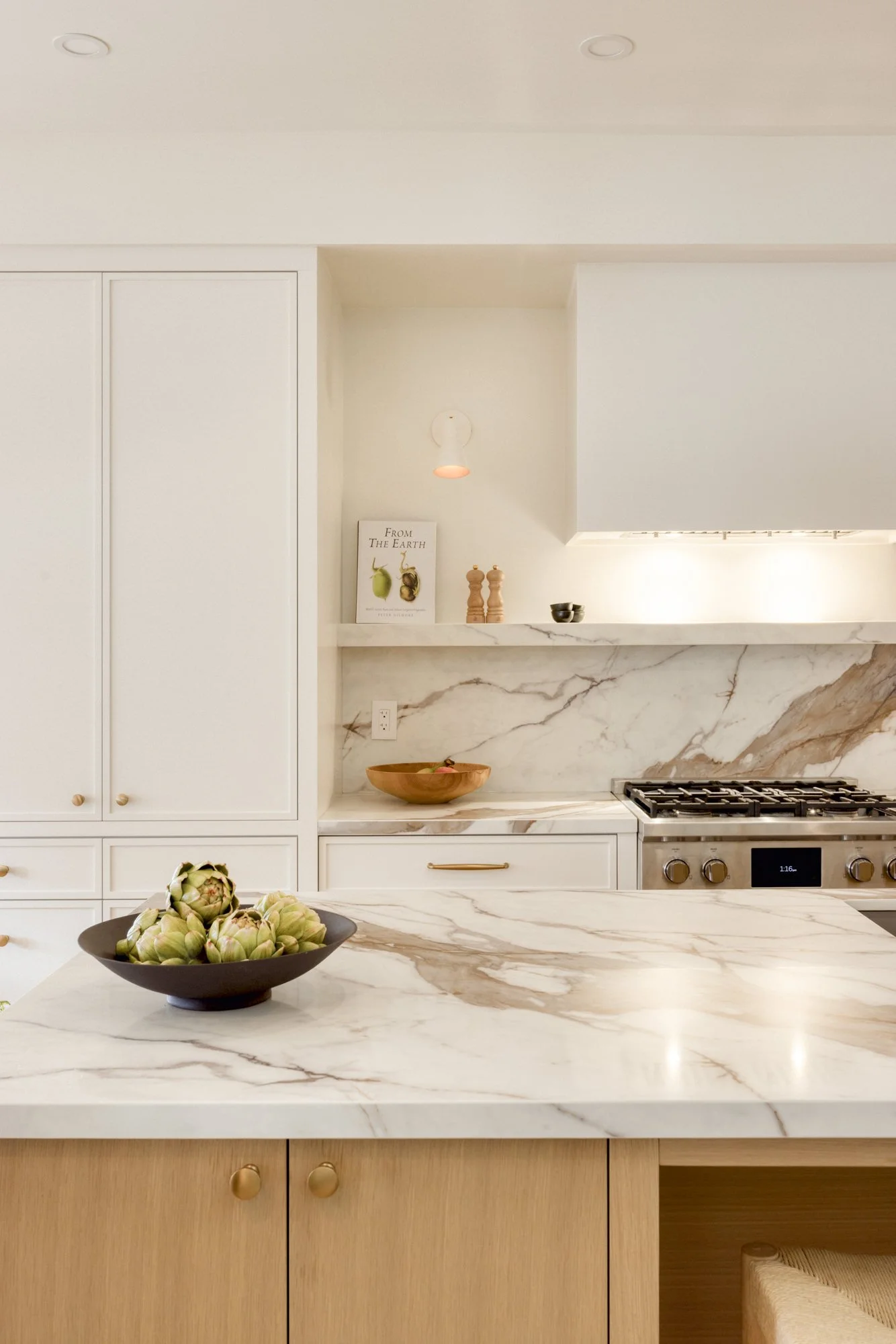 A close-up of a modern kitchen with white marble countertops, a black bowl of artichokes, and cream-colored cabinets with gold knobs.