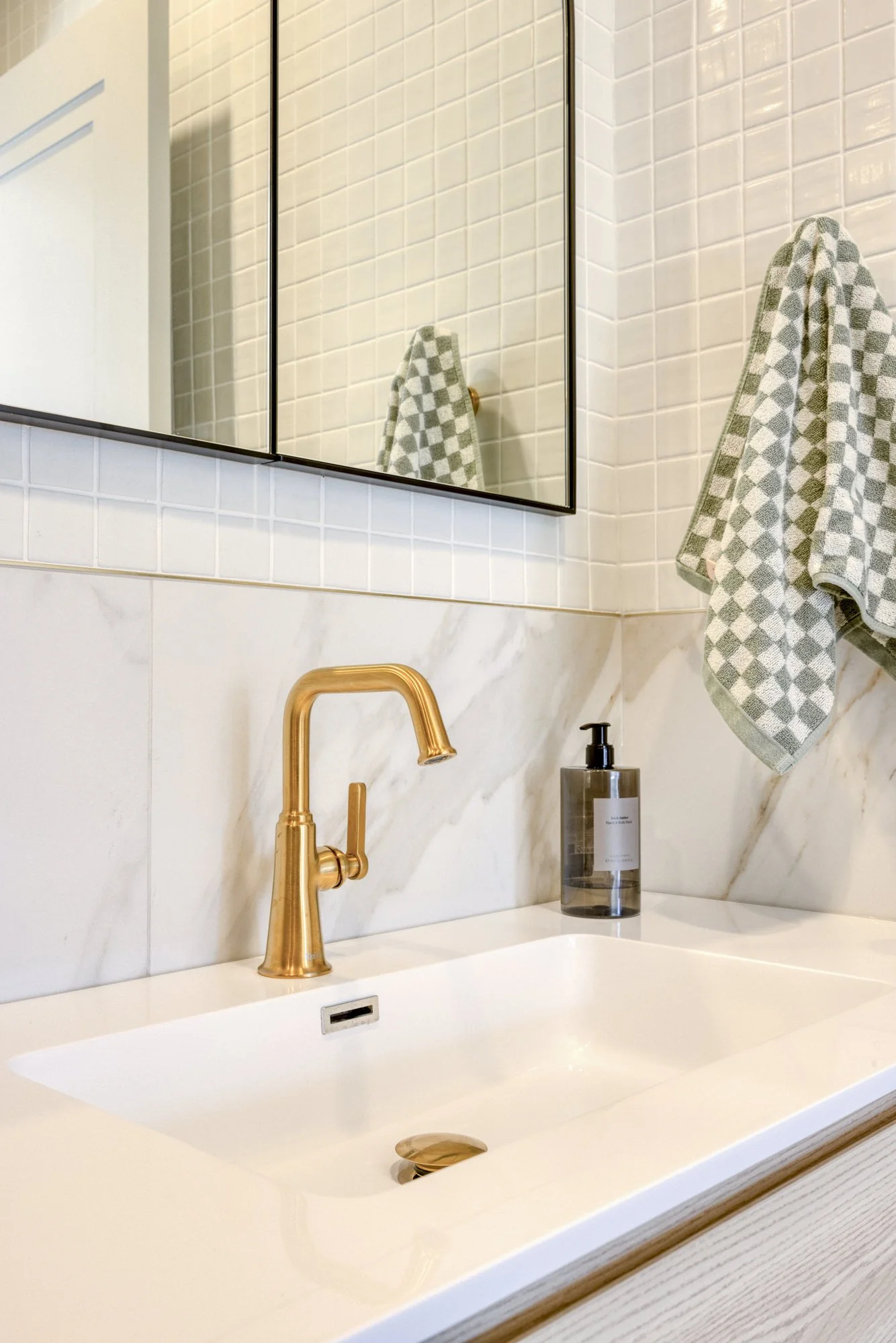 Bathroom sink with gold faucet, soap dispenser, mirror, and checkered towel on wall.