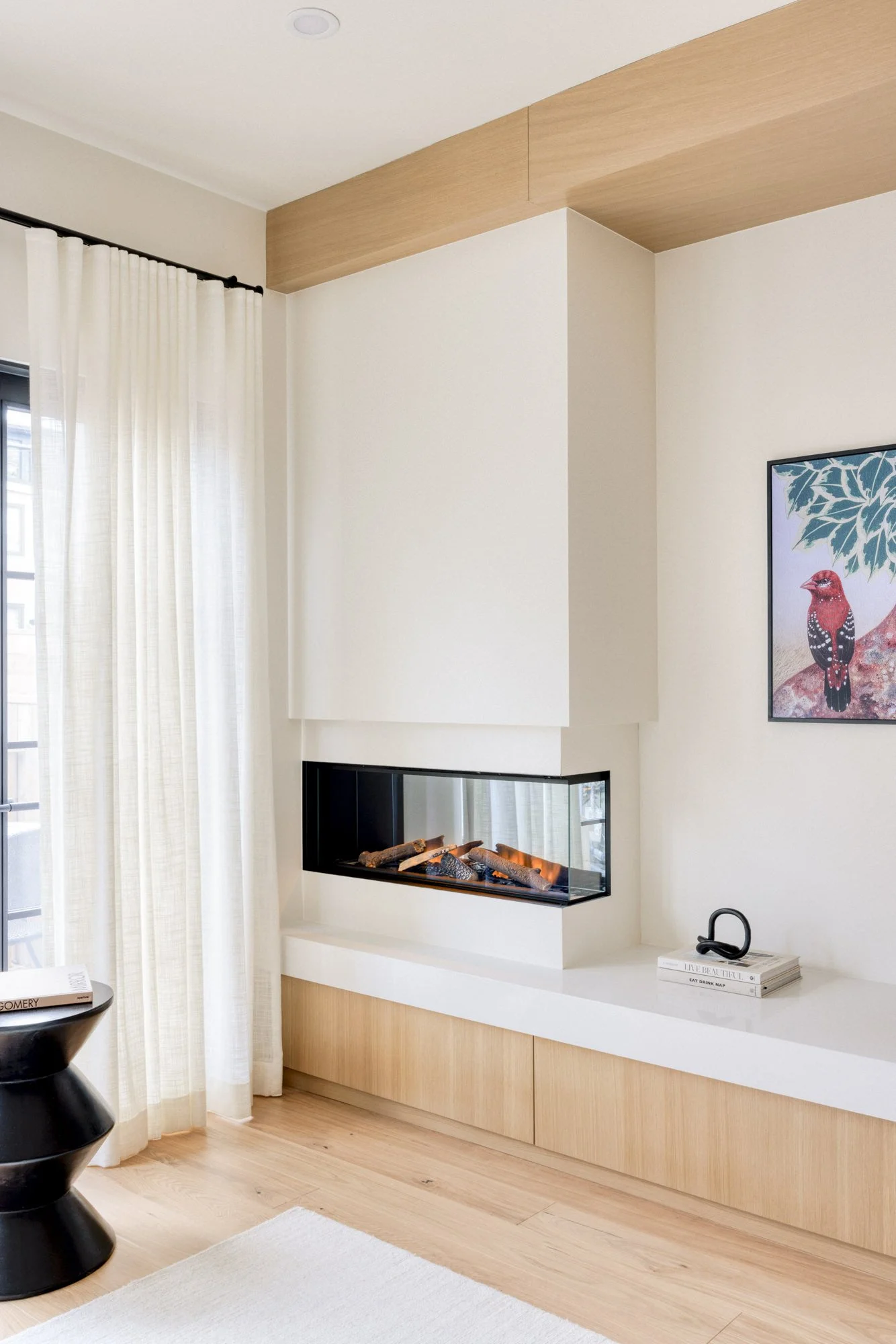 Modern living room with a corner fireplace, light wood flooring, white curtains, a black accent table, and minimalist decor including a book, headset, and colorful bird artwork on the wall.