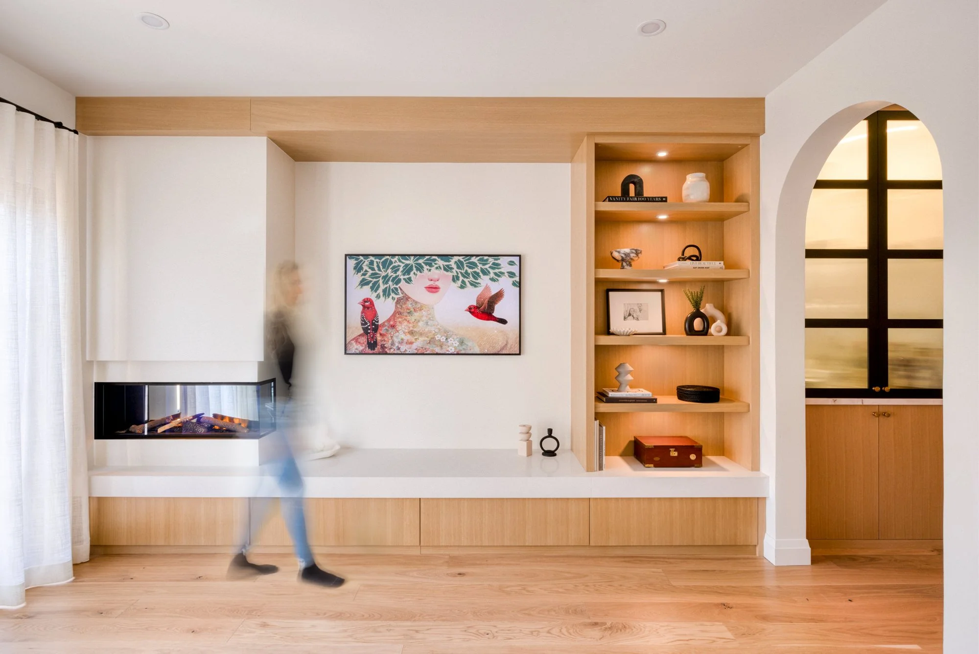 Modern living room with a built-in wooden shelf, a framed art piece of a woman's face with bird illustrations, a white fireplace, and blurred person walking by near a window with white curtains.
