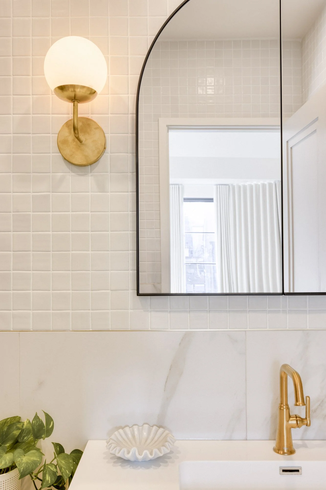 Bathroom with a large mirror, gold wall sconce, plant, decorative bowl, and gold faucet.
