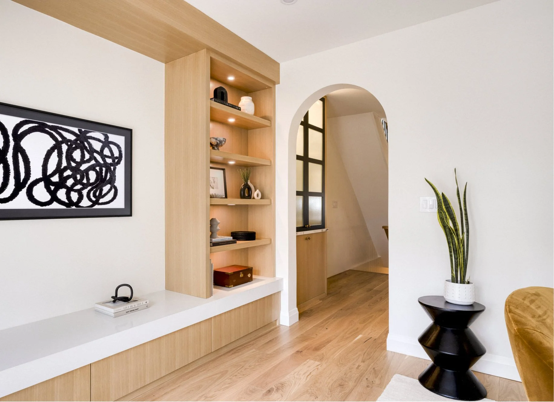 Modern living room with wooden flooring, a built-in shelf with decorative items, a framed abstract art, and a large houseplant in a white pot on a black side table.