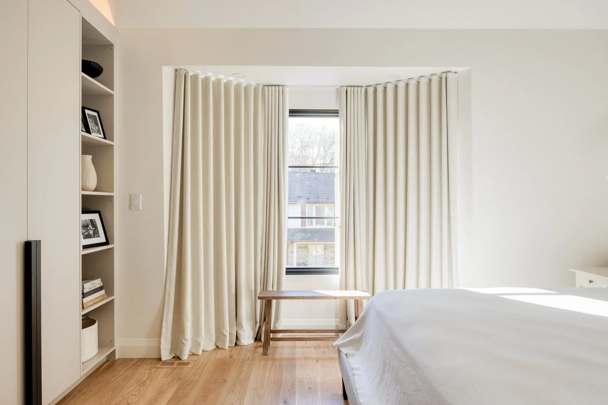 Bright bedroom with large window, cream curtains, wooden floor, white bed, and built-in shelves with black-and-white photos and decorative objects.