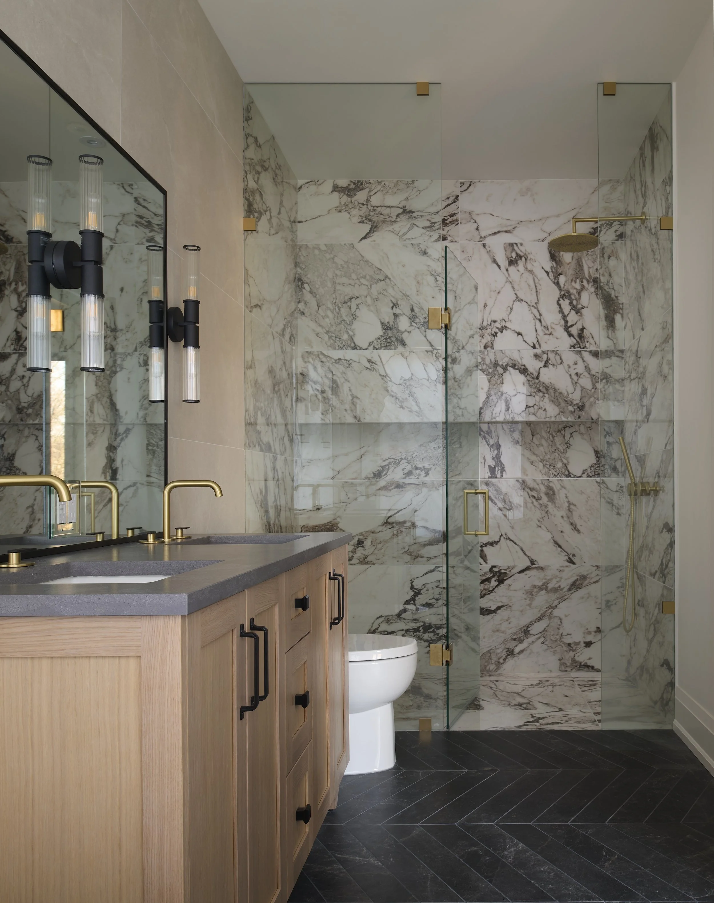 A Toronto interior by Design Works Studio: Modern bathroom with marble shower, wooden vanity with black handles, gold fixtures, and black herringbone floor.