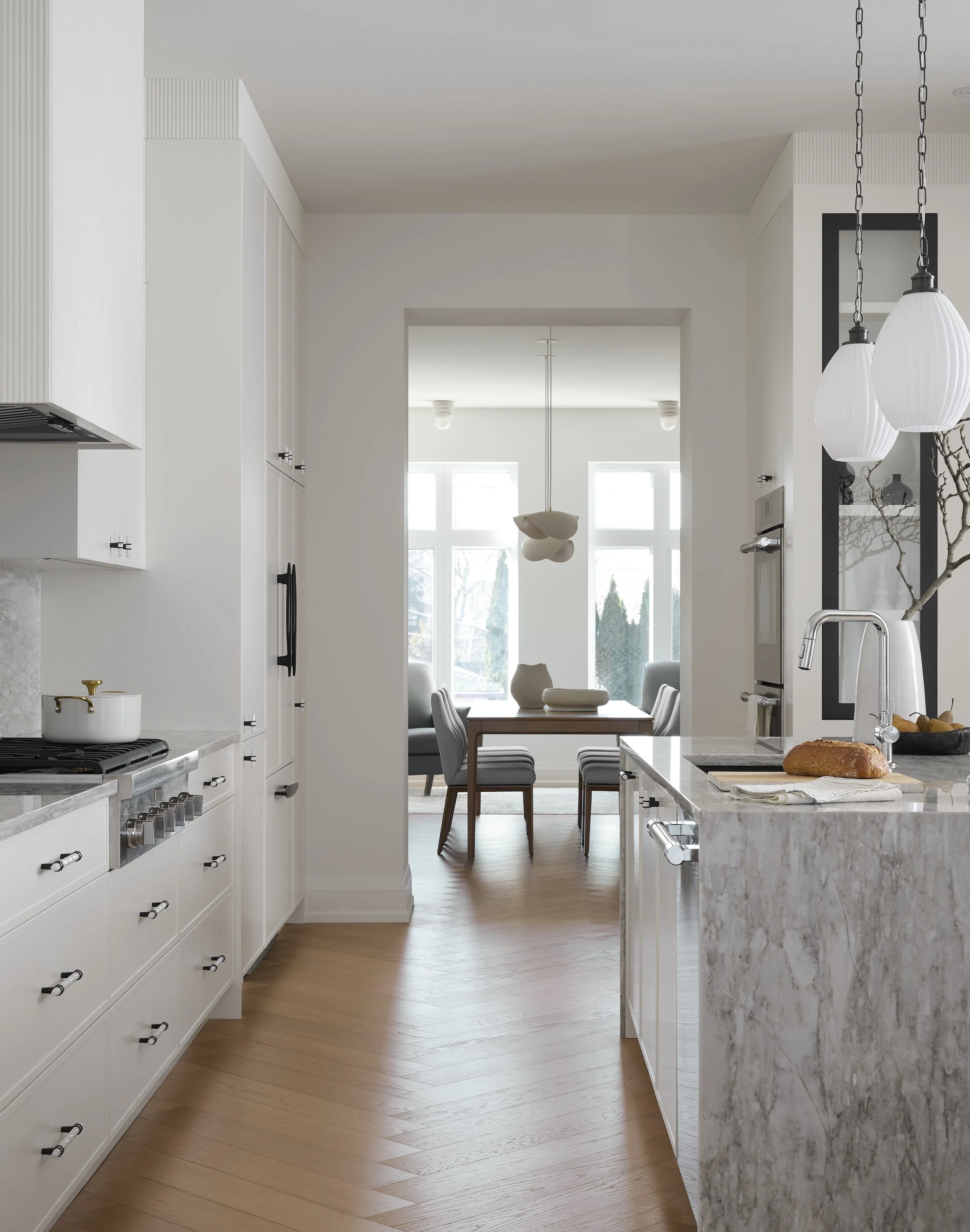 A Toronto interior by Design Works Studio: Modern kitchen with white cabinetry, marble countertop, and hardwood floors, leading into a dining area with large windows, a wooden table, and gray upholstered chairs.