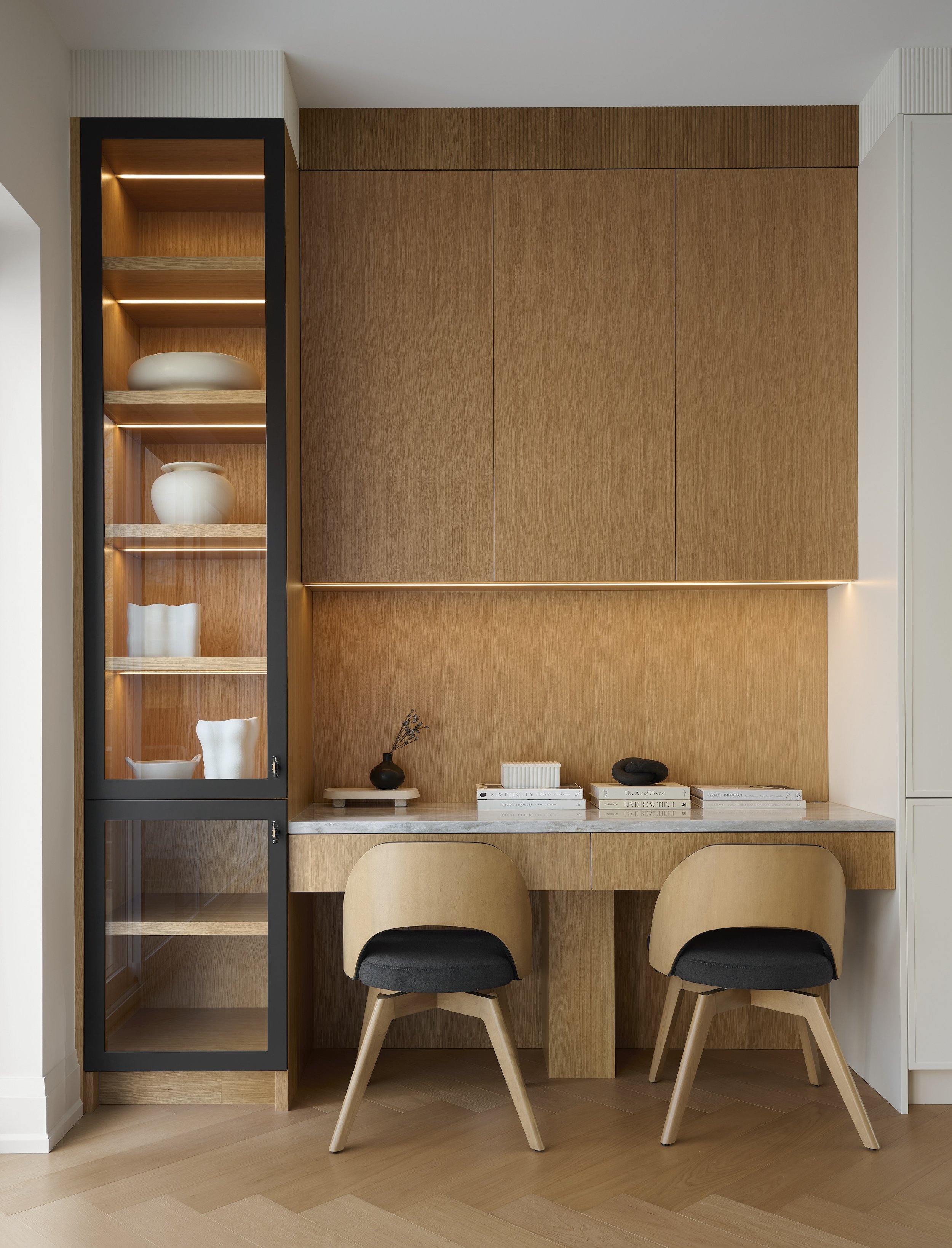 A Toronto interior by Design Works Studio: A minimalist wooden workspace with a built-in desk, two modern chairs, and a glass-front cabinet with decorative vases and bowls.