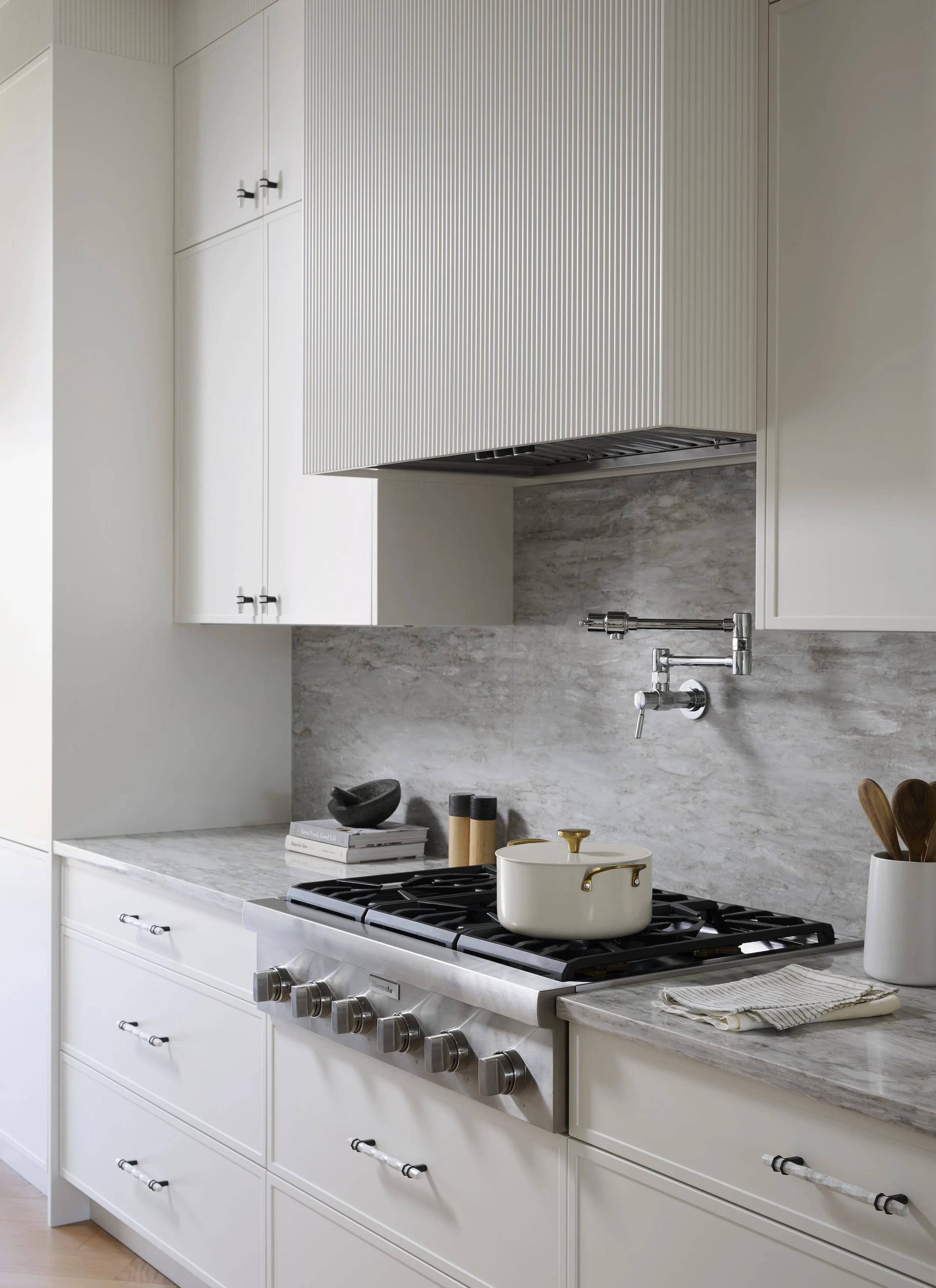 A Toronto interior by Design Works Studio: Modern kitchen with white cabinets, marble countertop, and a gas stove with a white pot. There are kitchen utensils and books on the counter, with a textured backsplash and a built-in range hood.