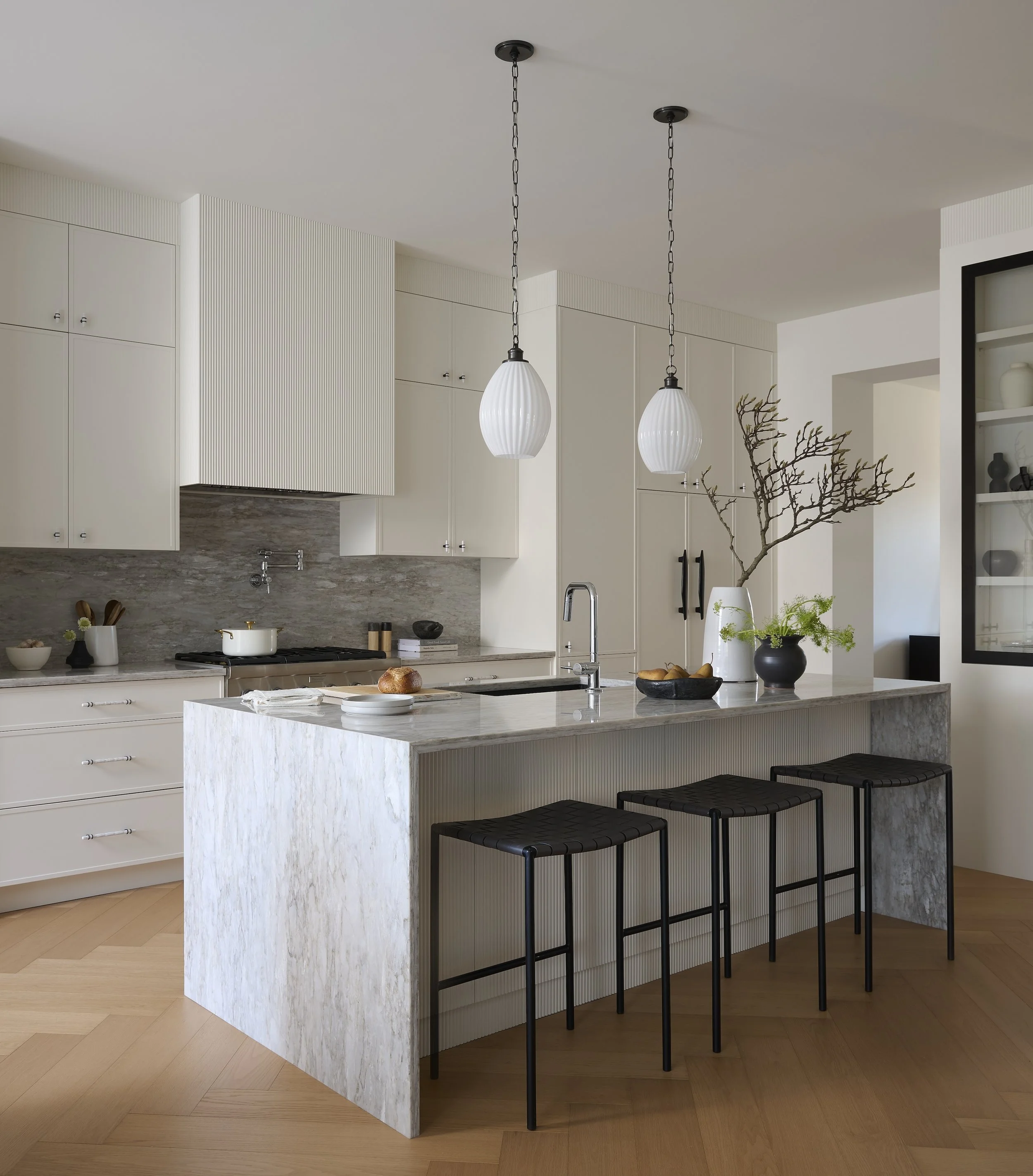 A Toronto interior by Design Works Studio: Modern kitchen with white cabinets, marble island, black bar stools, pendant lights, and decorative plants.