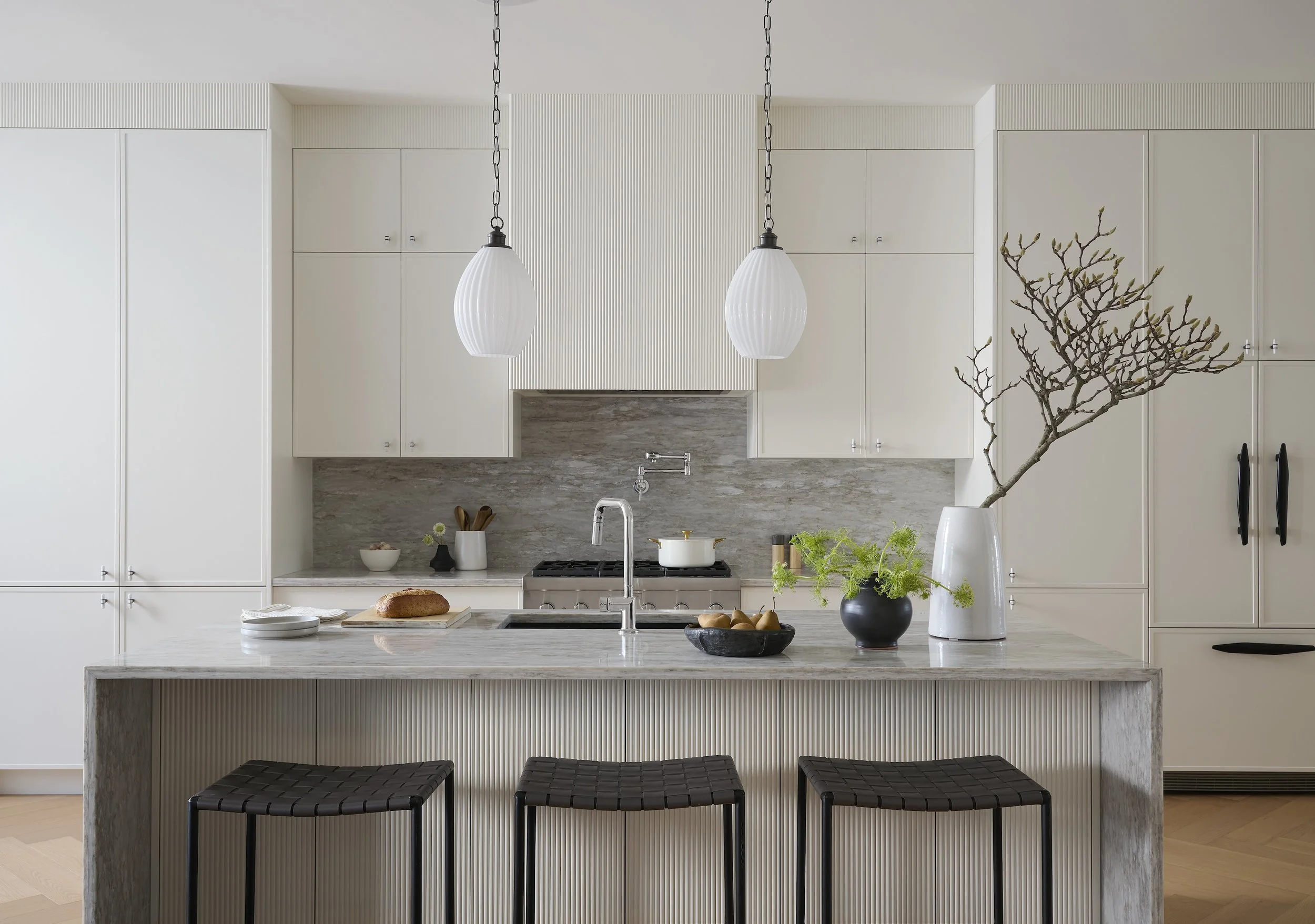A Toronto interior by Design Works Studio: Modern kitchen with white cabinetry, gray marble backsplash, and a marble kitchen island with black woven barstools. Decor includes a large white vase with branches.