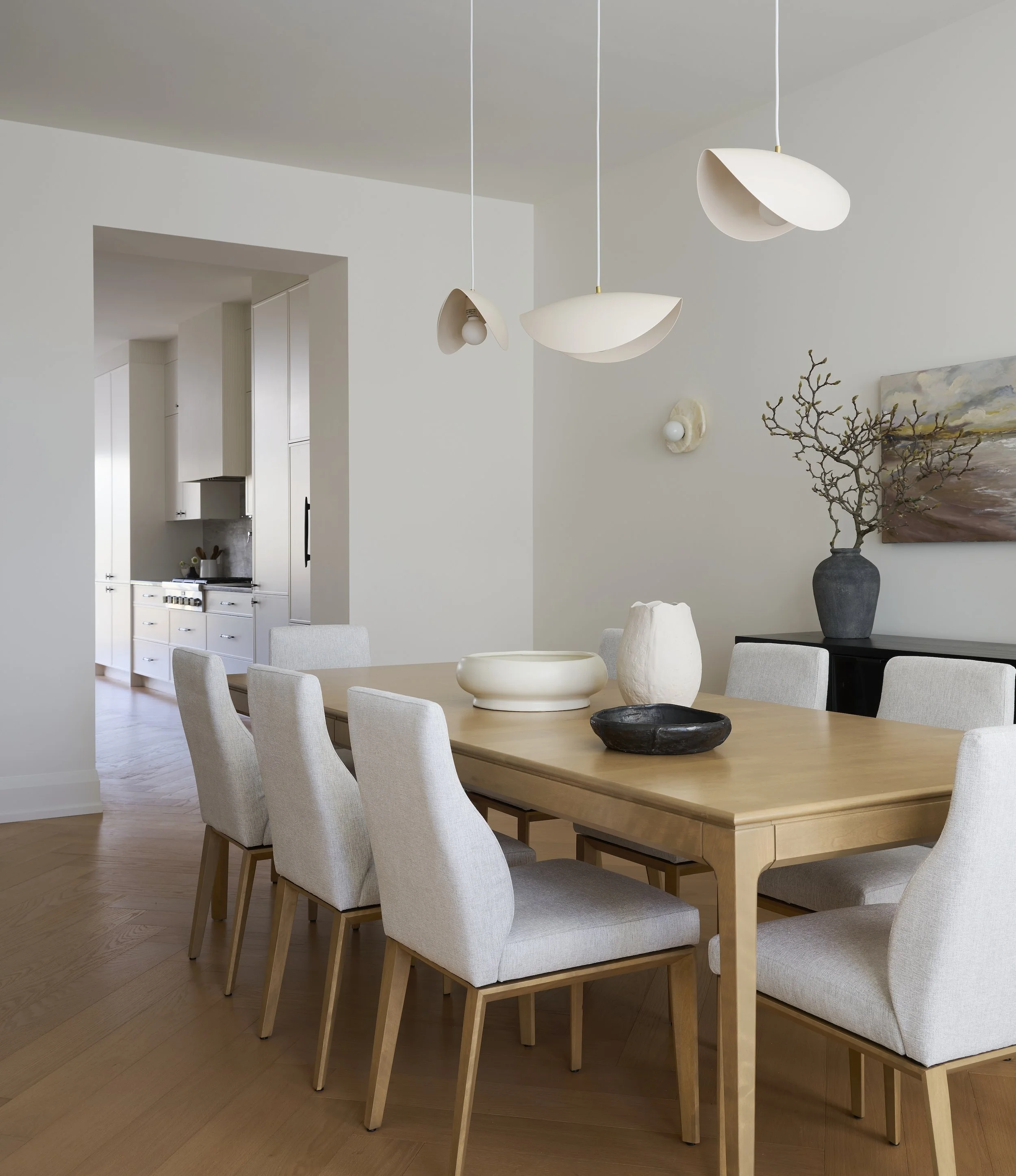 A Toronto interior by Design Works Studio: Modern dining room with a wooden table, white upholstered chairs, decorative vases, and abstract wall art, with pendant lighting overhead.
