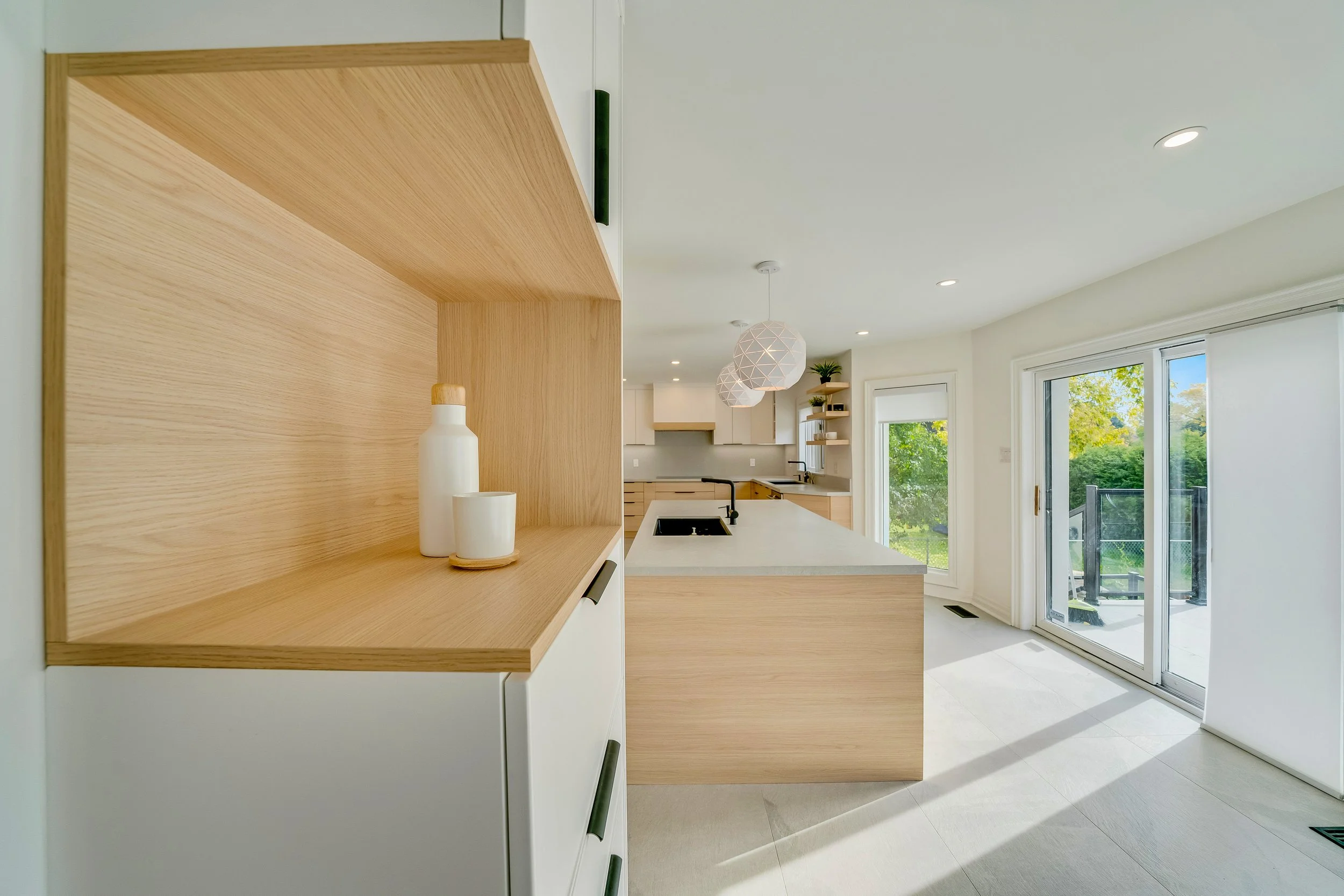 Bright, modern kitchen with wooden cabinets and white countertops, featuring a sliding glass door leading to an outdoor balcony with a view of green trees.