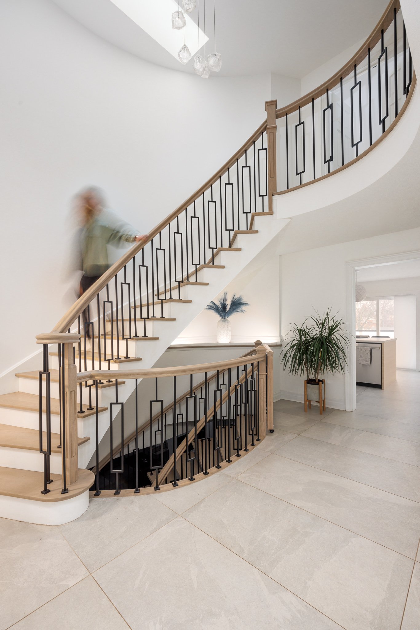 Interior view of a modern staircase with light wood handrails and black metal balusters, beside white walls and potted plants, with a blurred person walking down the stairs.