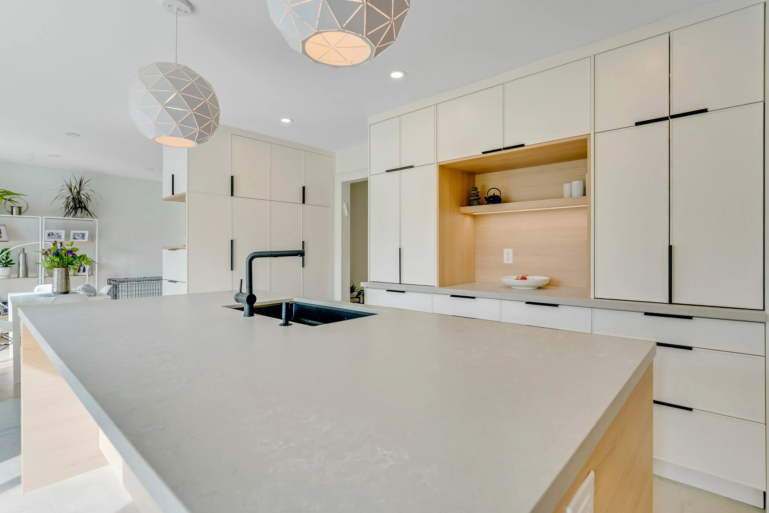 Modern kitchen with white cabinets, a large white countertop island with a black sink and faucet, and minimalist decor including a bowl of fruit and small decorative items. Pendant lights hang from the ceiling, and a glimpse of the dining area and li